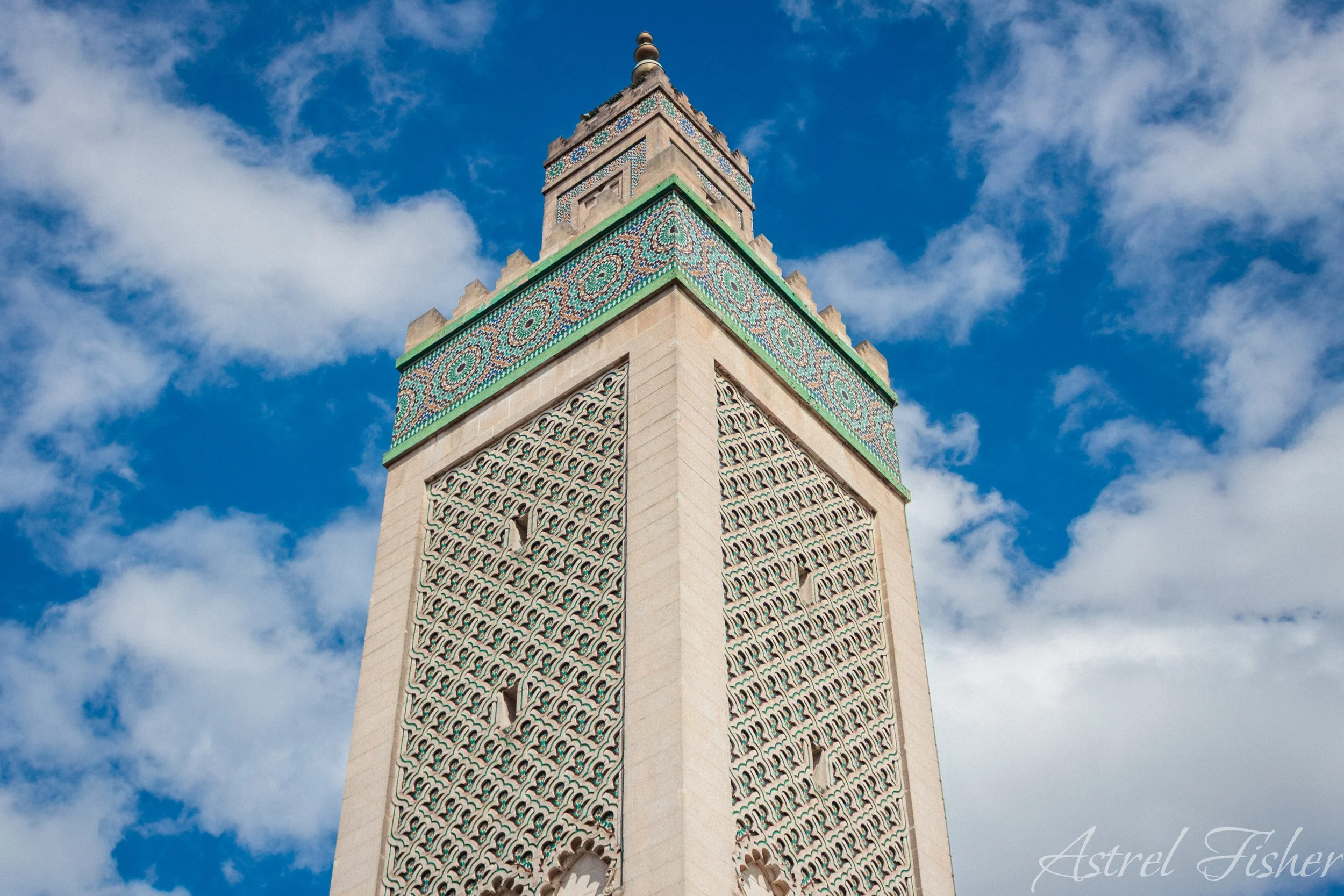 A tall minaret with intricate turquoise and white tile patterns near the top, set against a blue sky with scattered clouds.