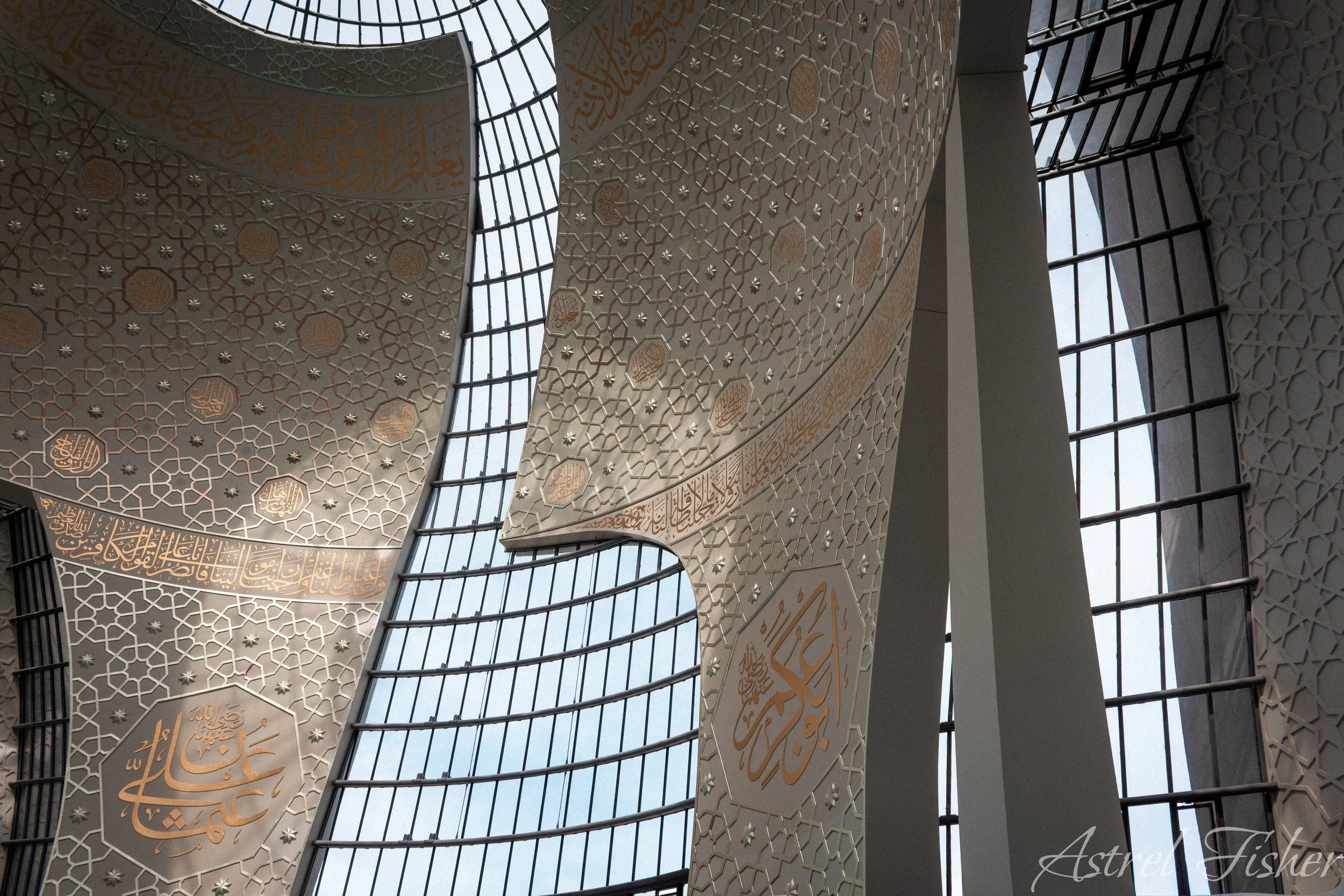 Interior view of a mosque ceiling with intricate Islamic calligraphy and geometric patterns in gold on white panels, framed by large glass windows with black metal bars.
