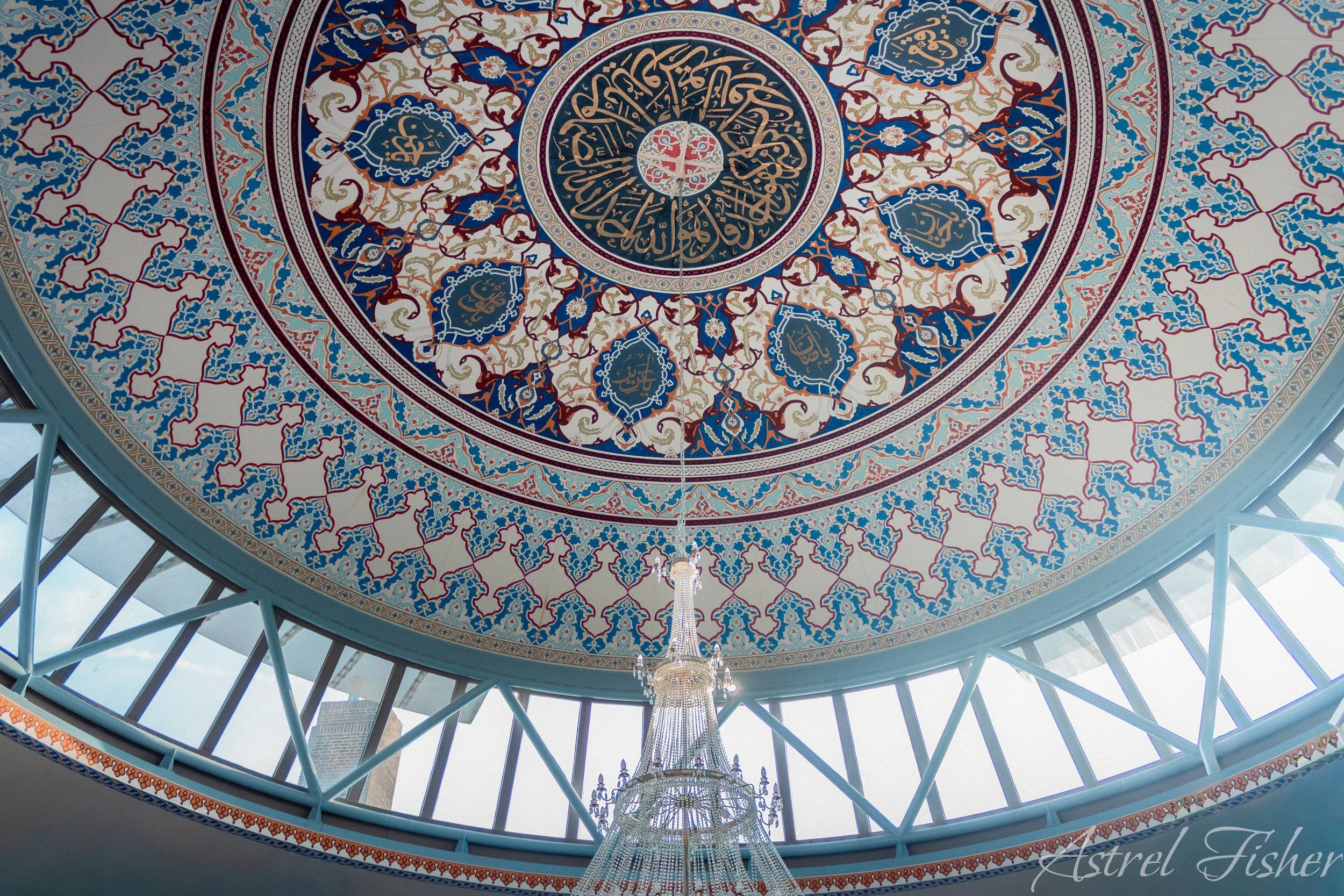 Interior view of a domed ceiling with intricate Islamic geometric patterns, calligraphy, and a chandelier hanging in the center, seen through a glass roof with metal framework.