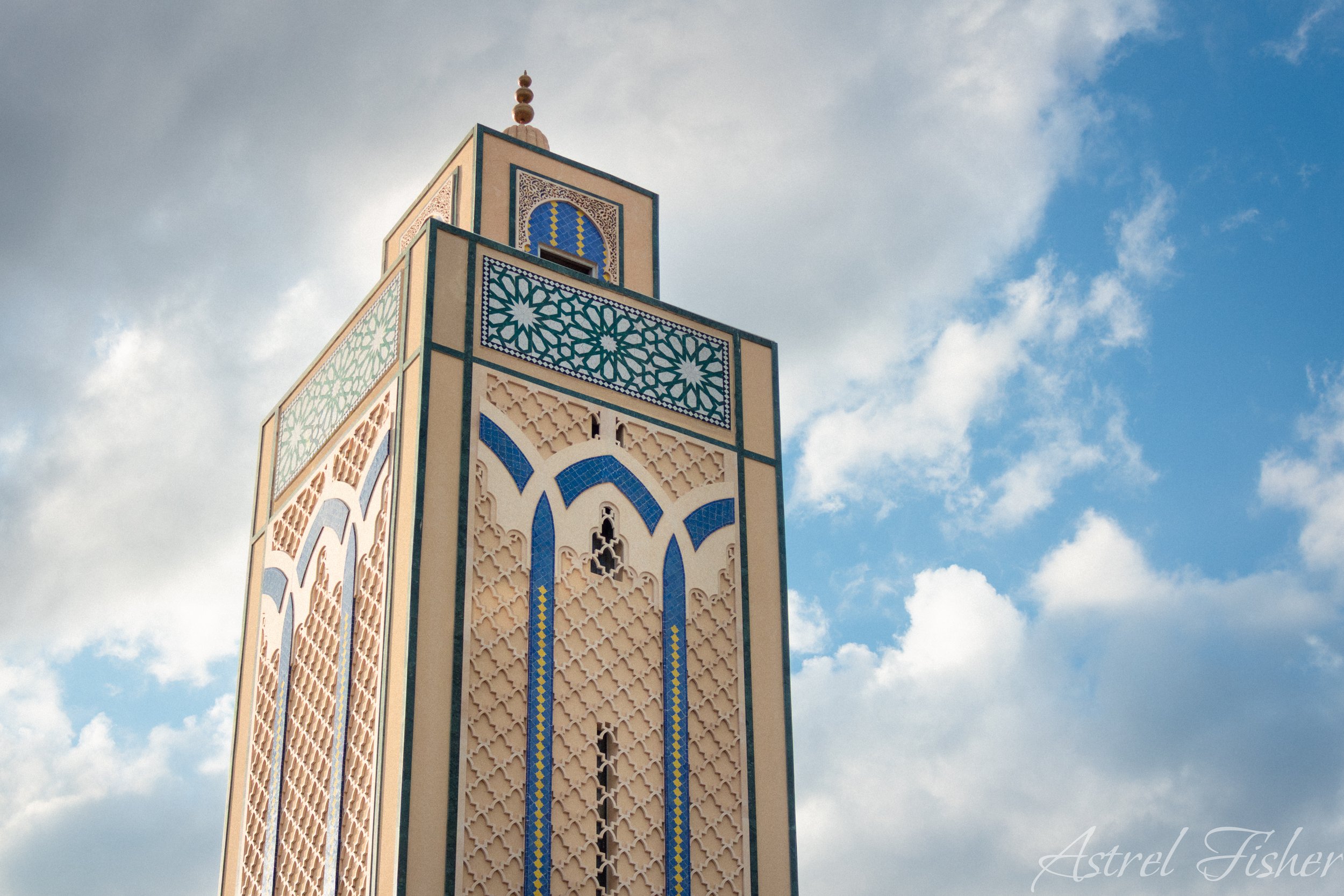 A close-up of a tall minaret with intricate tile work and geometric patterns in beige, blue, and green colors, set against a partly cloudy sky.