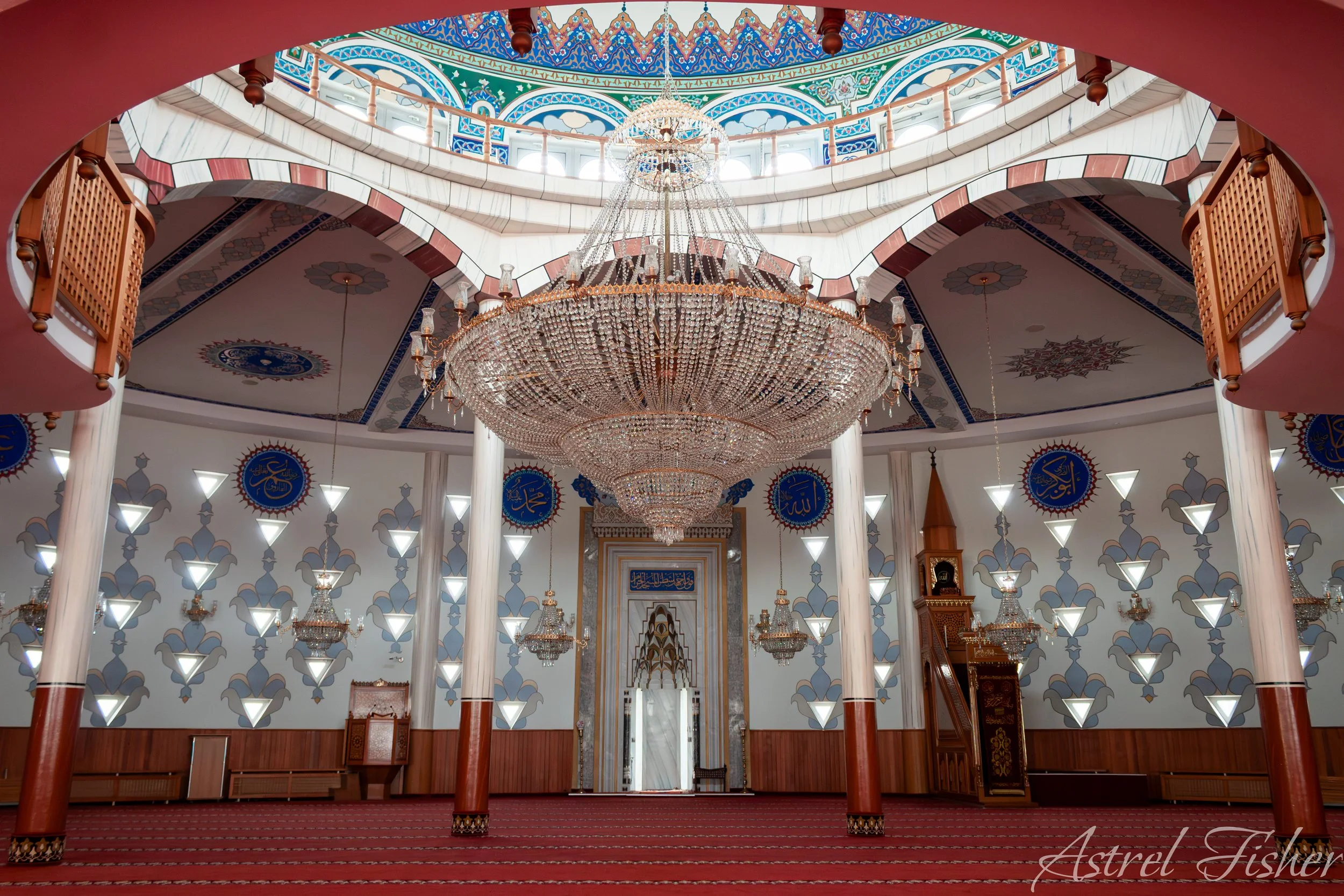 Interior view of a mosque with a large central chandelier, decorated walls, and upper galleries with wooden railings, patterned arches, and Arabic calligraphy.