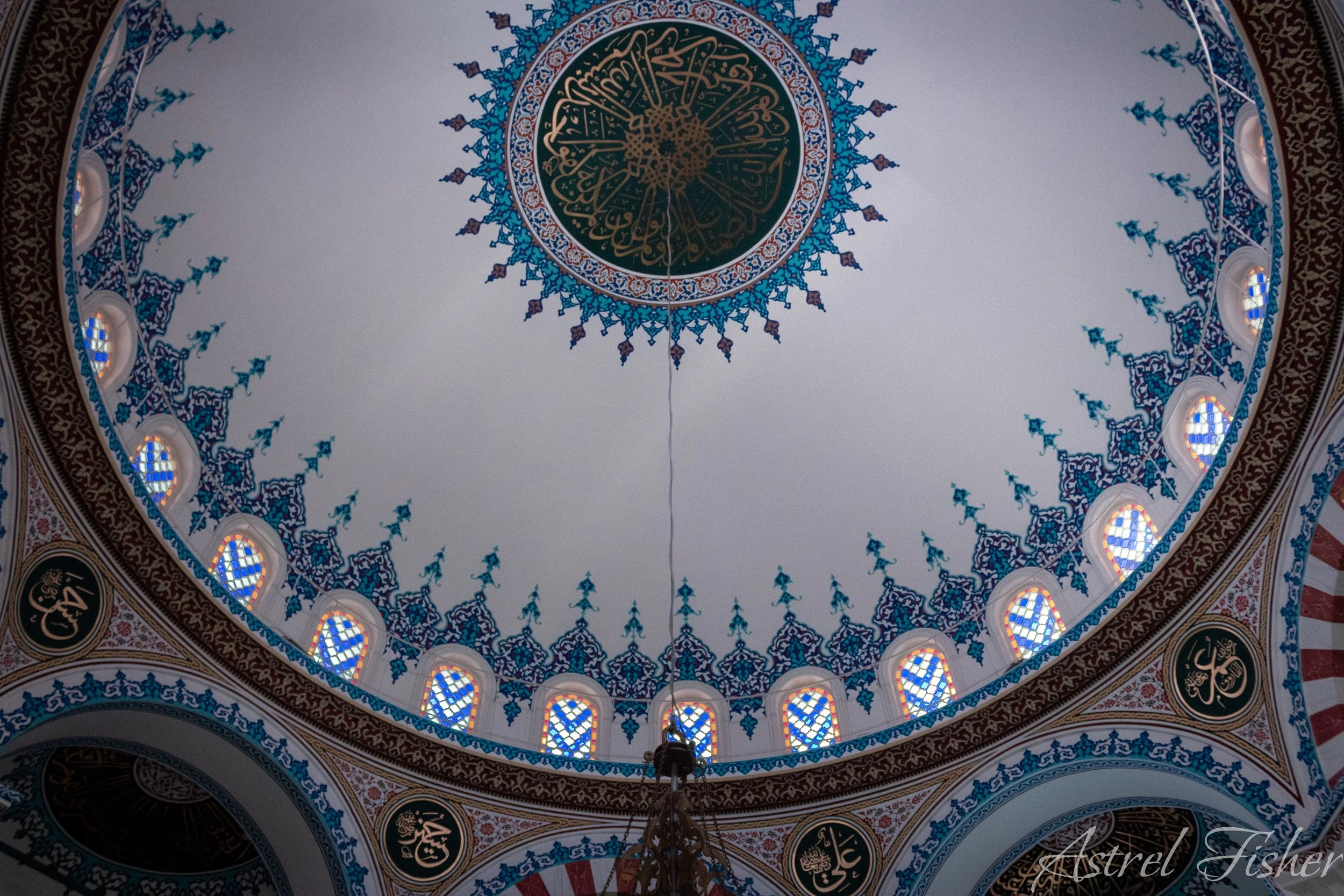 The ceiling of a mosque with intricate Islamic geometric and calligraphic designs, featuring a large circular central pattern with calligraphy surrounded by stained glass windows and ornate decor.