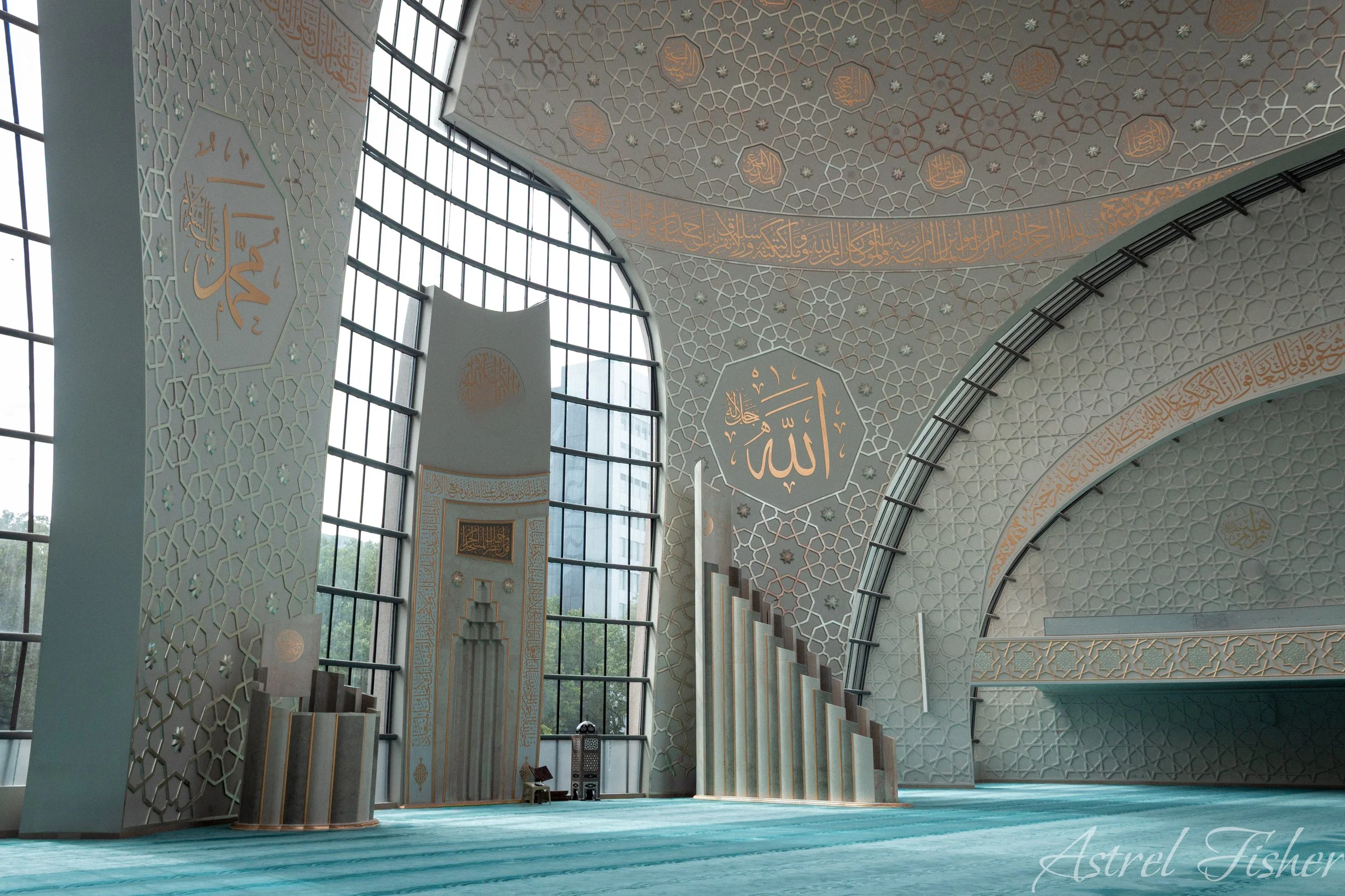 Interior of a mosque with intricate Islamic geometric patterns on the ceiling, large windows, and a prayer area with rows of prayer rugs.