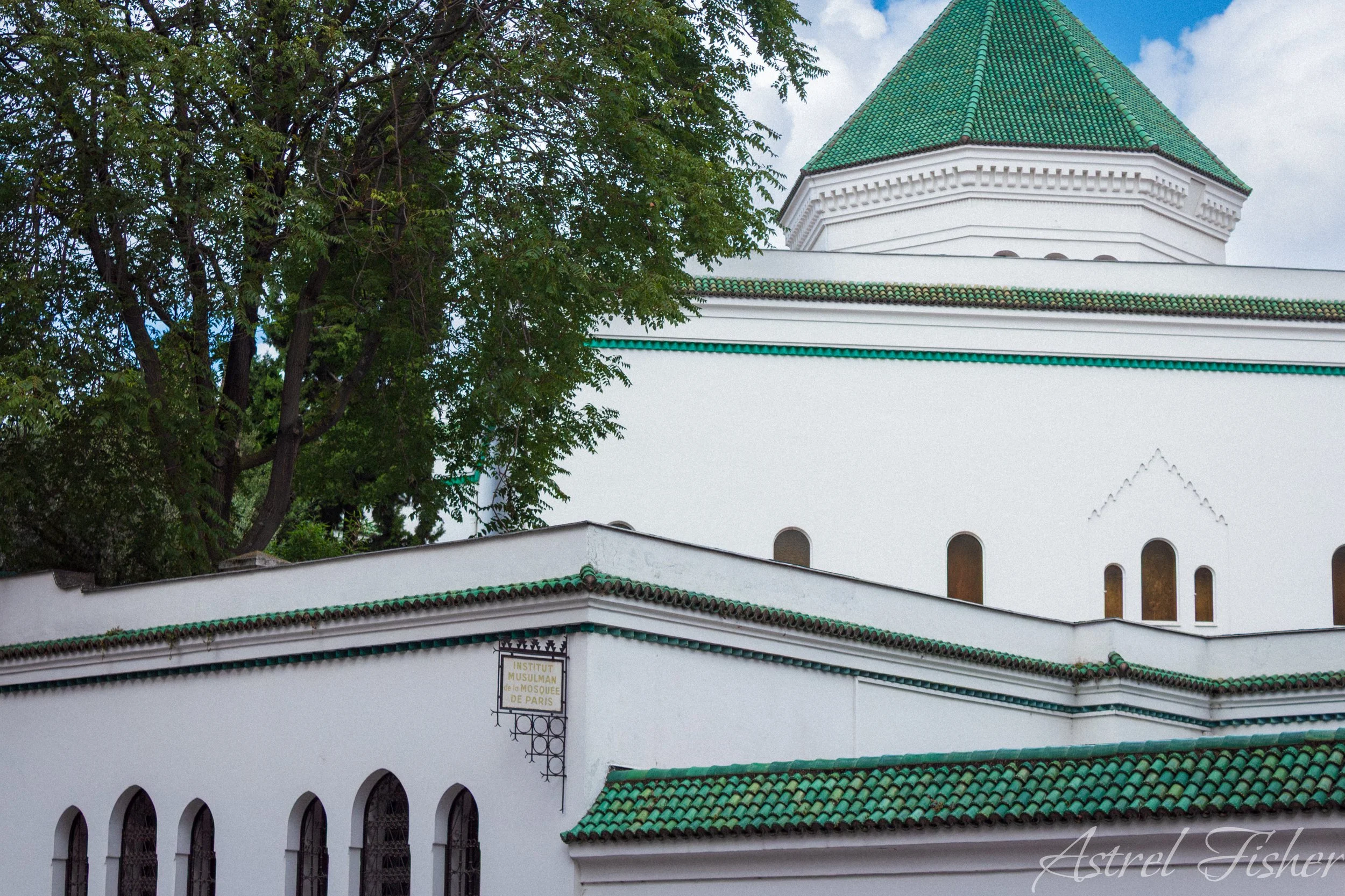 White building with green tiled roof, arched windows, and a sign indicating it is the Muslim Mosque of Paris, partially obscured by tree foliage.