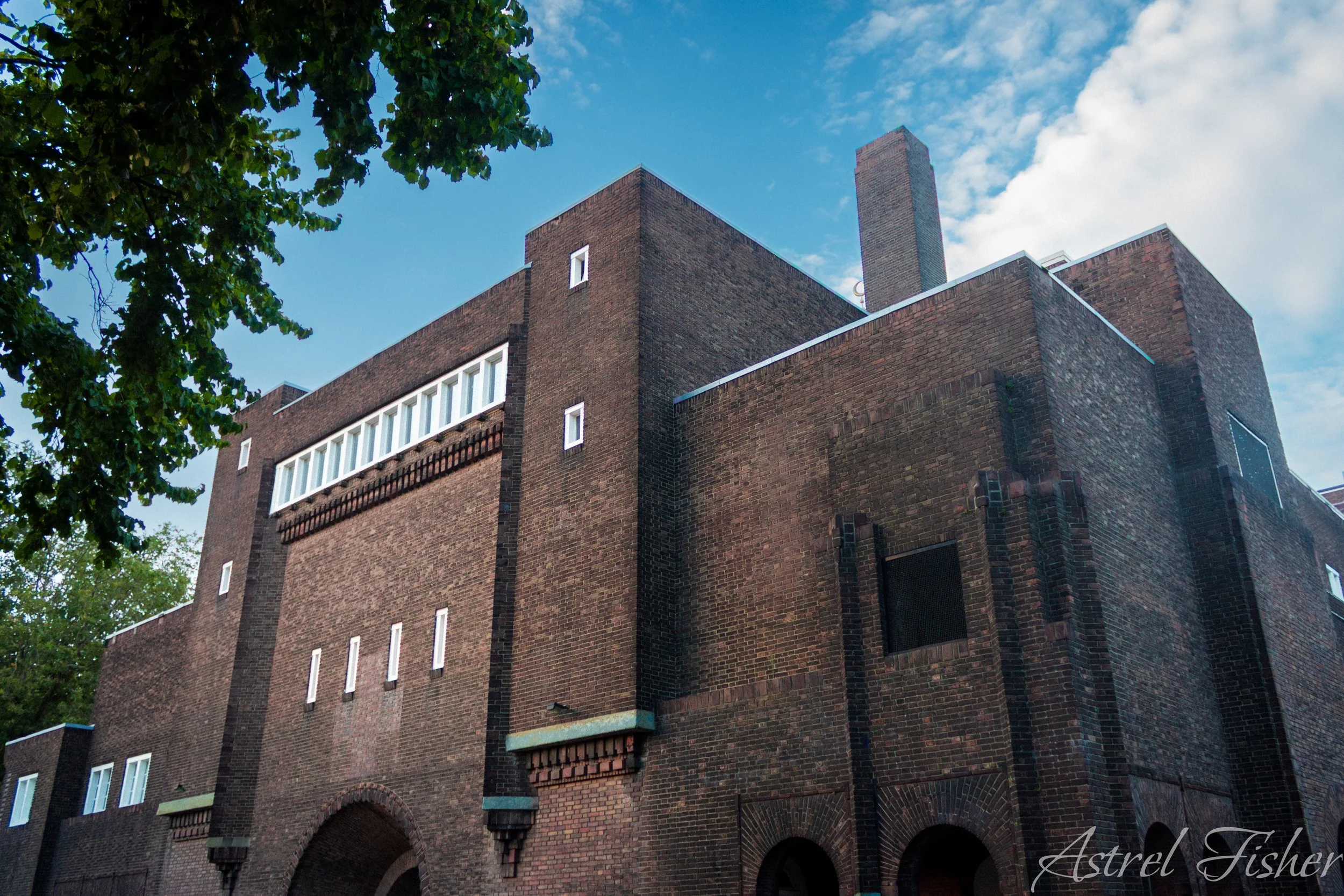 A large brick building with small windows and arched entrances at the bottom, under a bright blue sky with some clouds. Green tree leaves partially frame the top left corner.