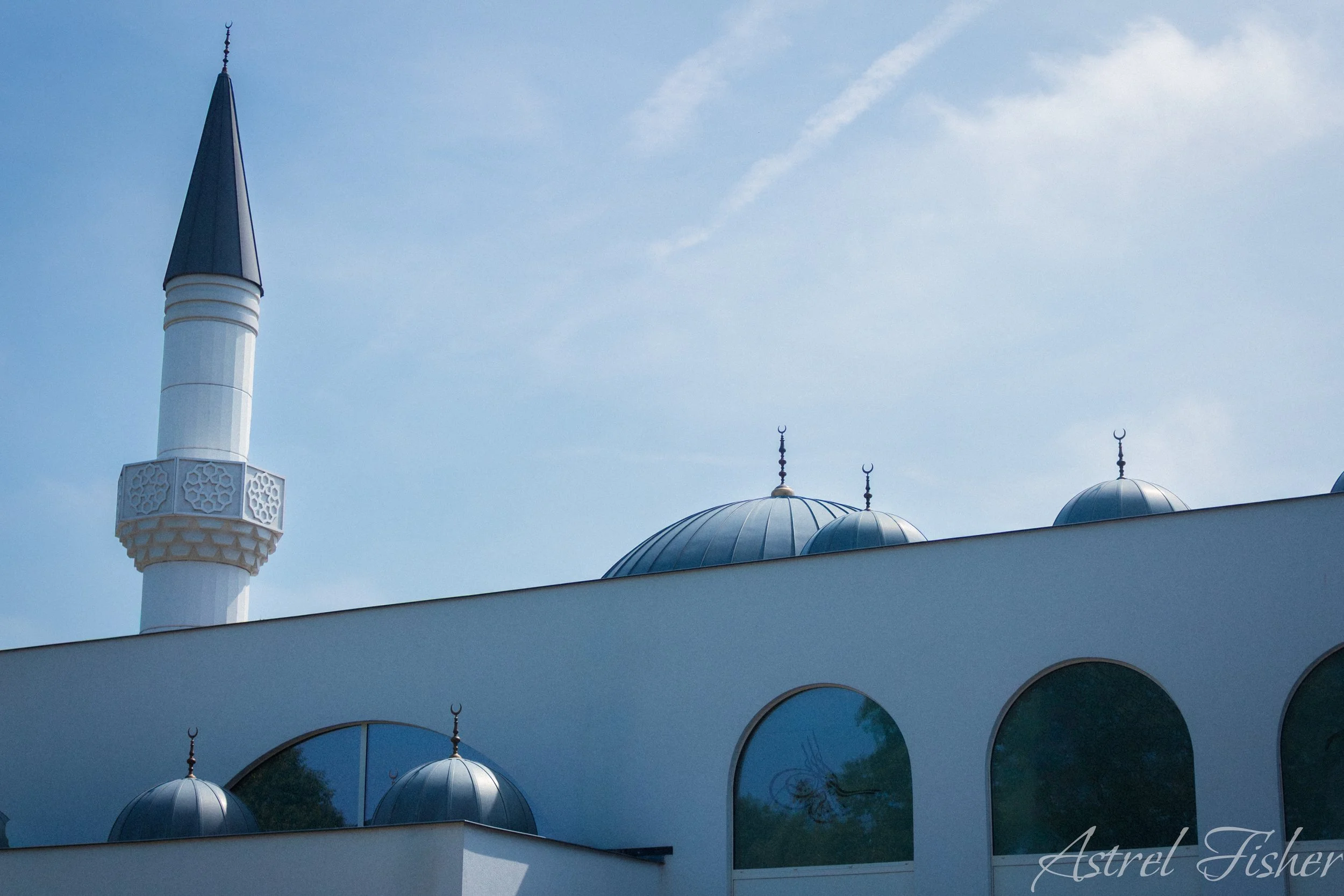 A white mosque with a tall minaret and multiple domes, against a blue sky with some clouds.