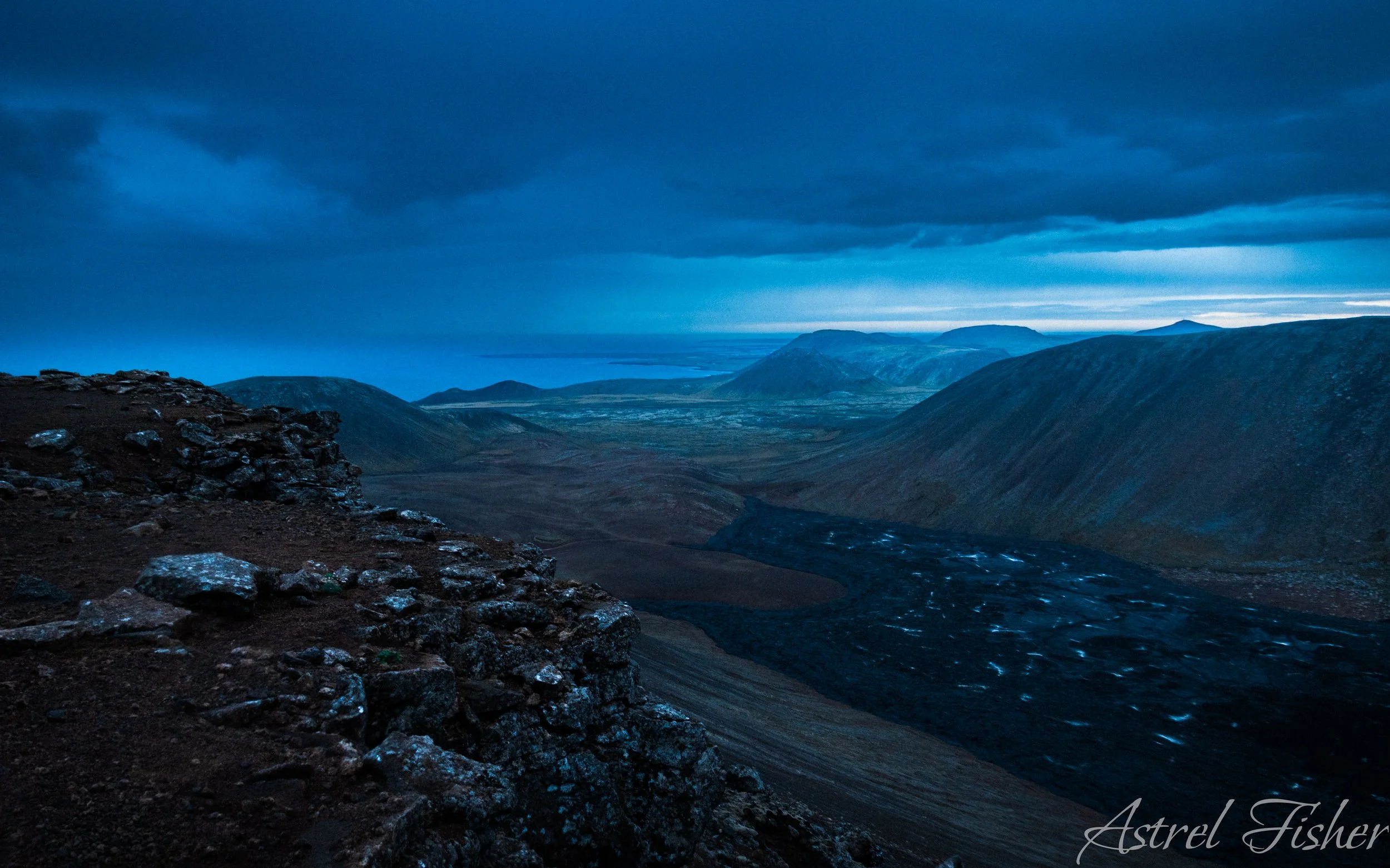 A rugged mountain landscape under a dark, cloudy sky with blue hues. Rocky terrain in the foreground and distant mountain ranges and valleys visible in the background.