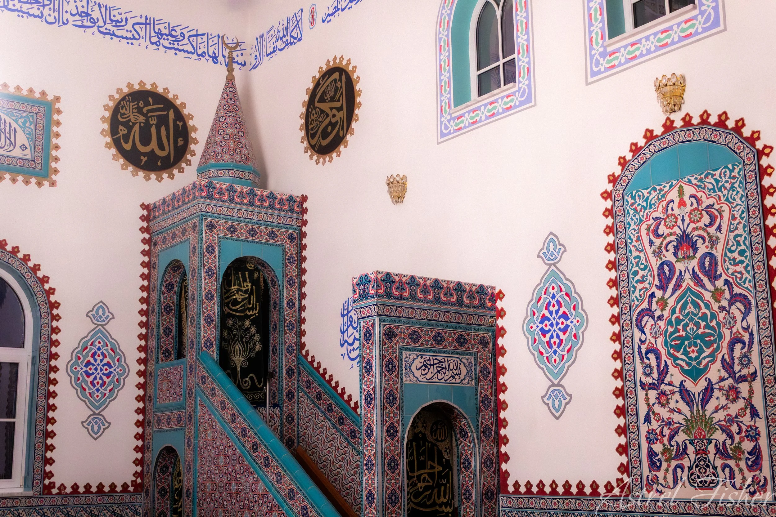 Interior of a mosque with intricate Islamic geometric and floral tile patterns in blue, red, and white, featuring display of Arabic calligraphy on walls and windows.
