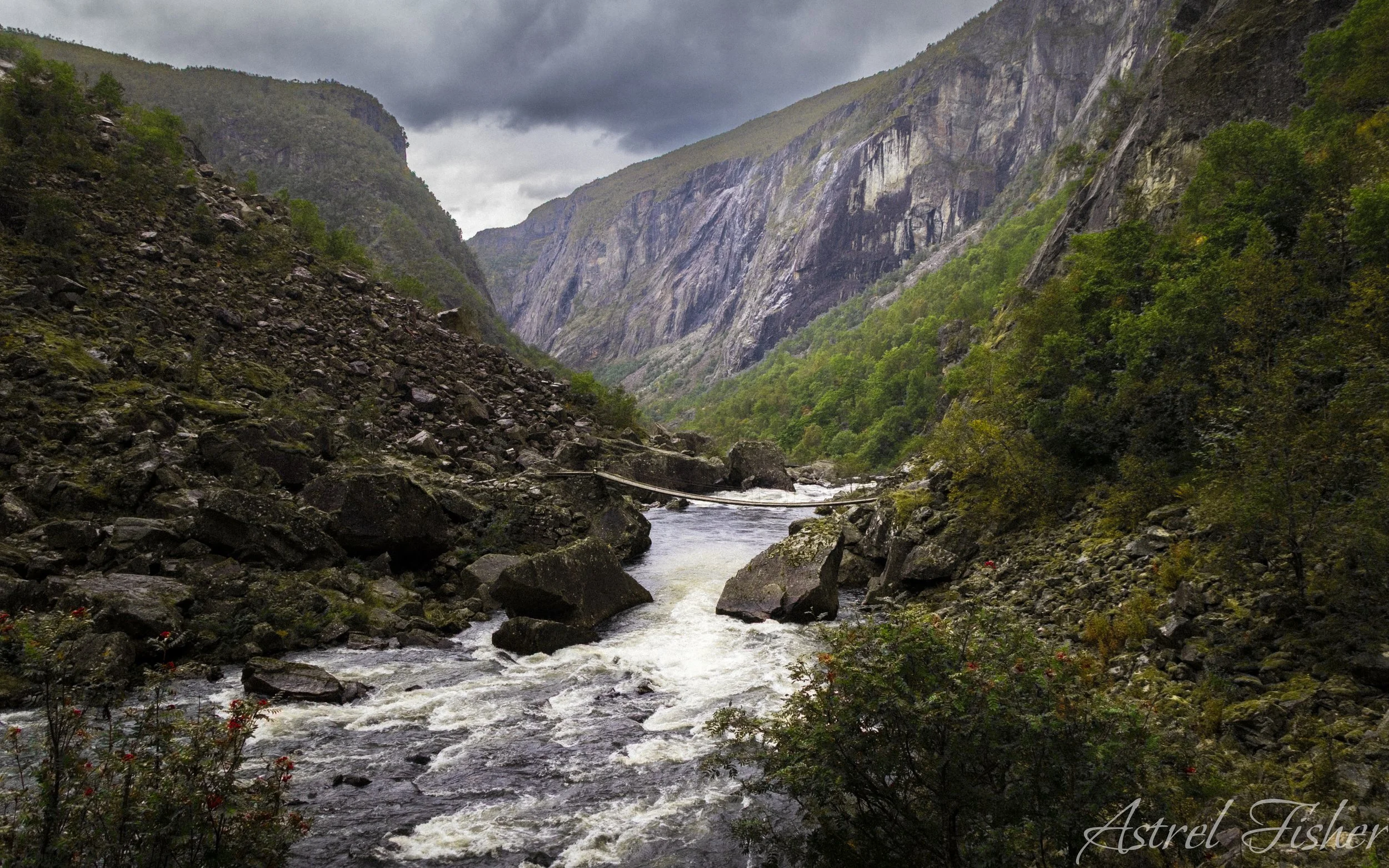 A mountain valley with steep, rocky, green-covered cliffs on both sides, a narrow river flowing through the bottom, and dark, cloudy skies overhead.