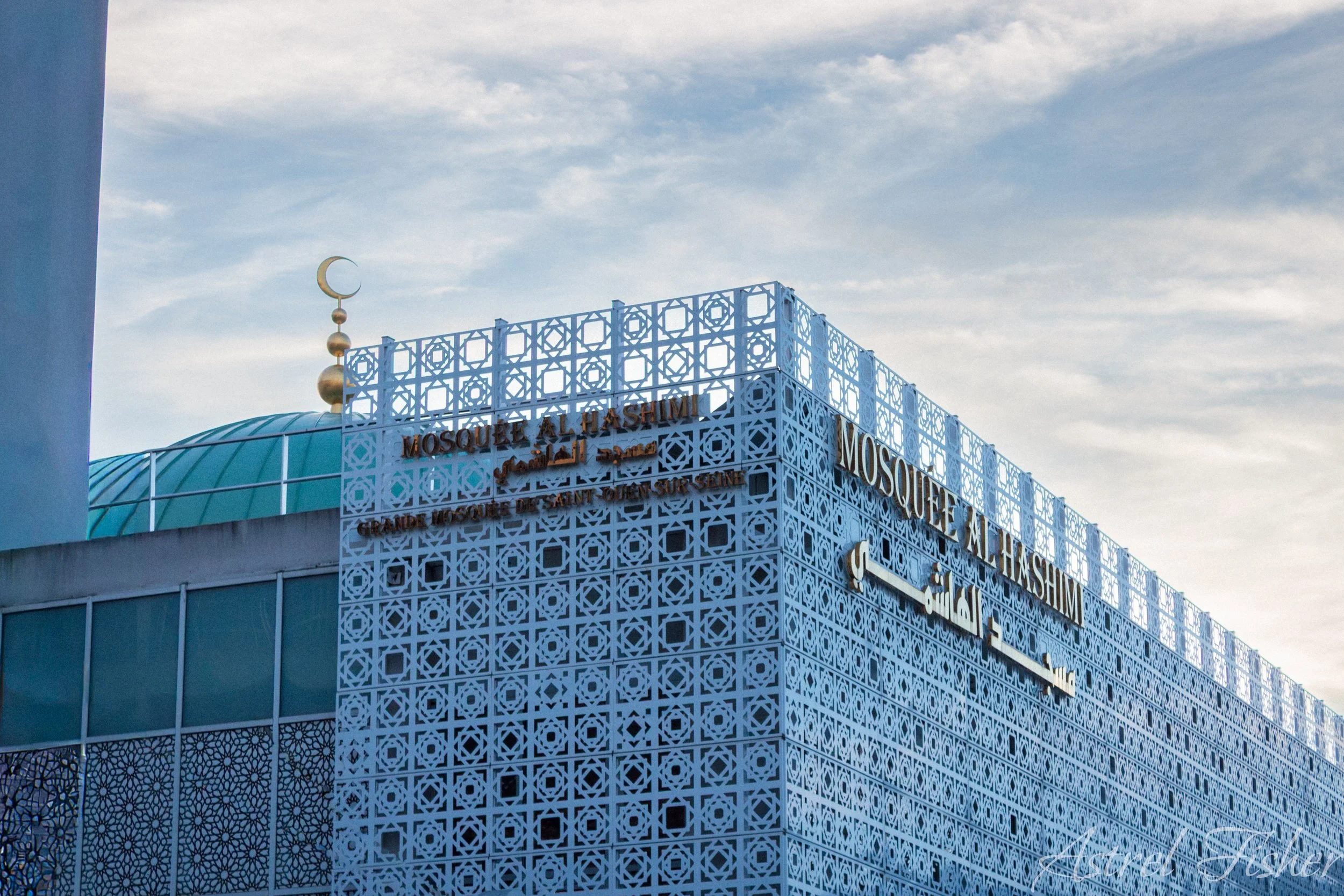 View of a mosque exterior with intricate geometric white latticework, a turquoise dome, and a crescent moon and star finial against a cloudy sky.