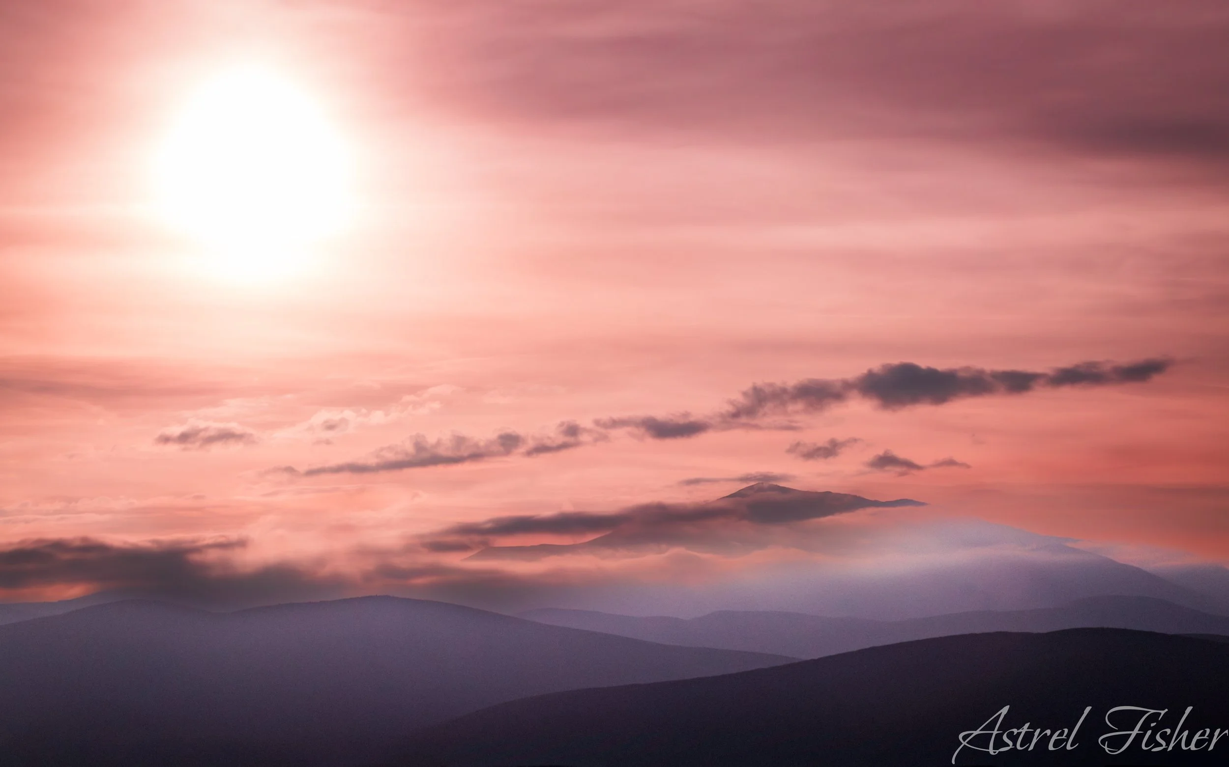 A scenic view of a sunset or sunrise over mountainous terrain with pink and purple sky, clouds, and a distant mountain peak.