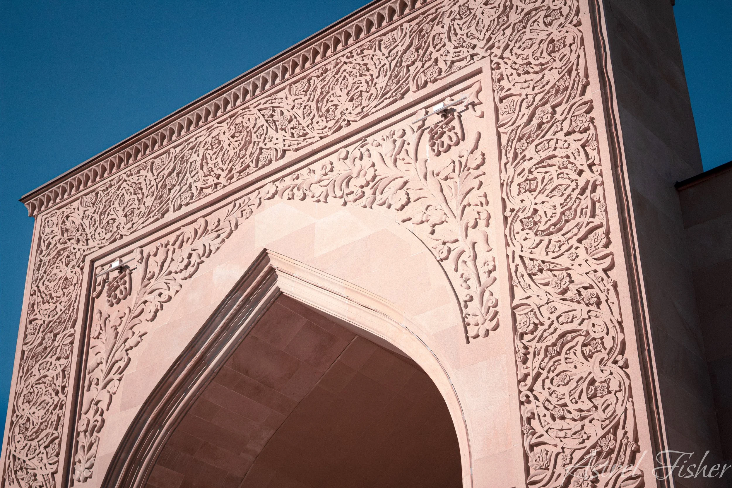 Close-up of a decorative archway with intricate floral and vine carvings in light pinkish stone against a blue sky.