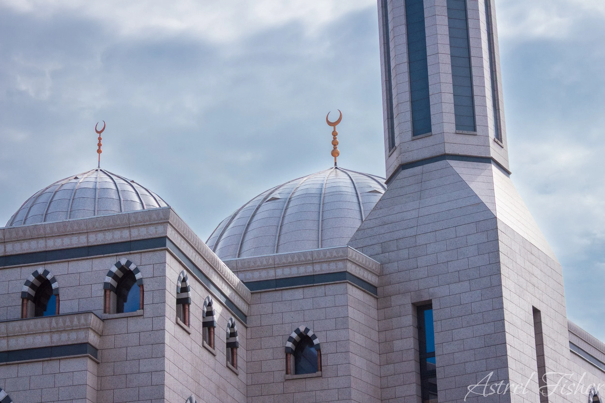 Close-up view of a mosque's domes and minaret against cloudy sky, with crescent moon finials on domes and minaret.