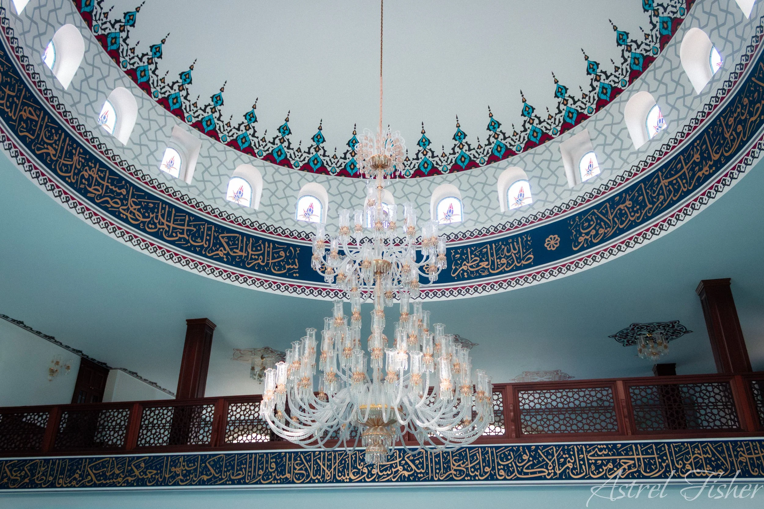 Inside a mosque with intricate Islamic calligraphy on the walls, decorative arch patterns, small windows in the upper wall, and a large chandelier hanging from the ceiling.