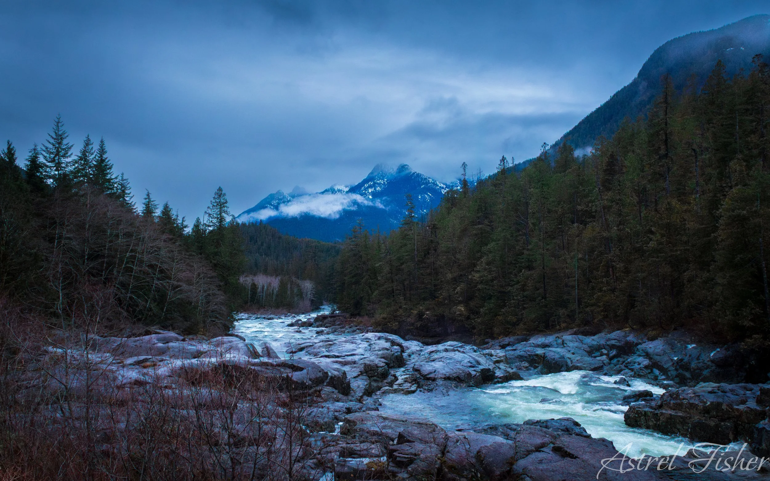A mountain landscape with snow-capped peaks in the background, a river flowing over rocks in the foreground, and a dense forest of conifer trees on either side.