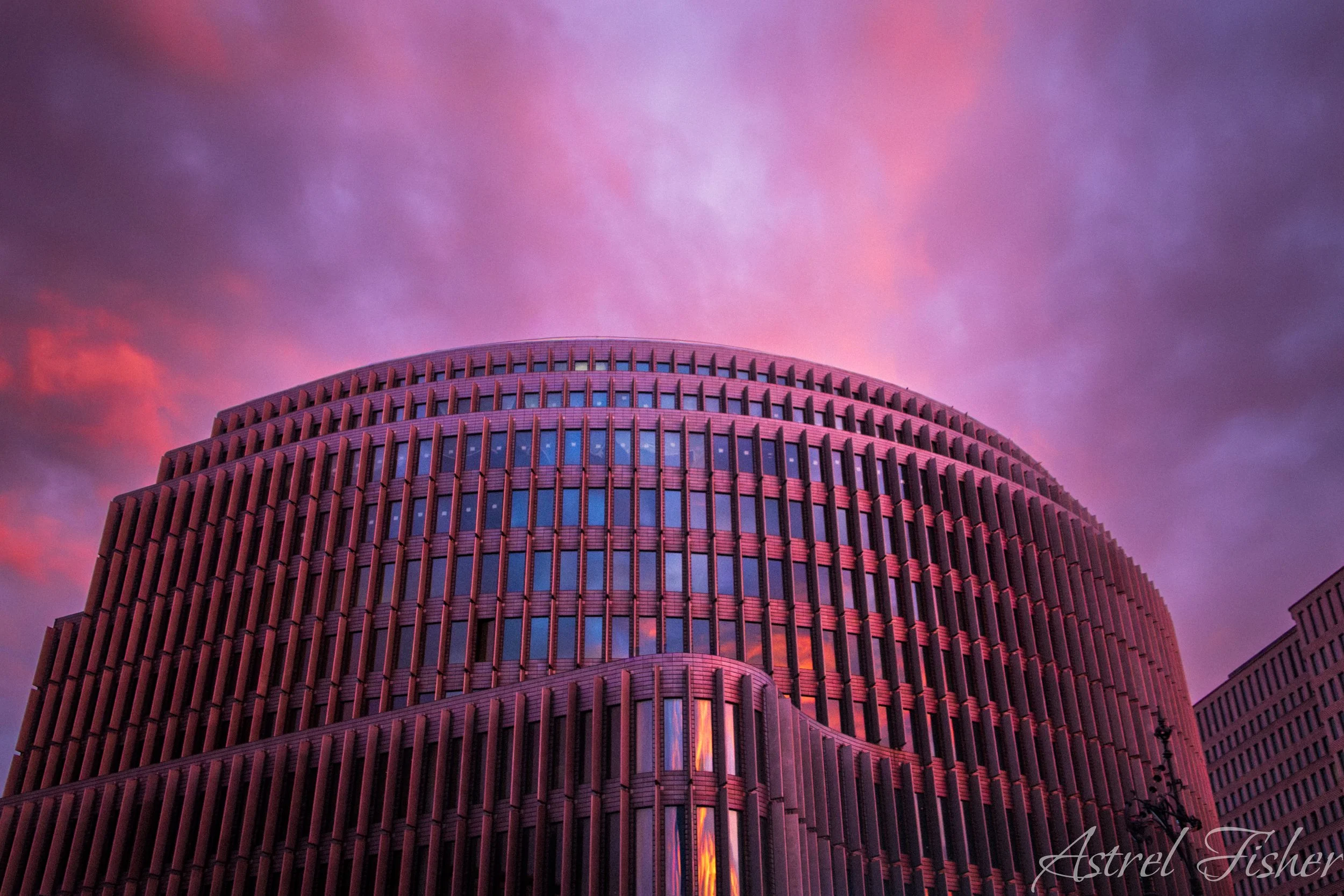 A modern, curved building with a grid of windows and vertical wooden slats, set against a colorful sky with pink, purple, and orange clouds.
