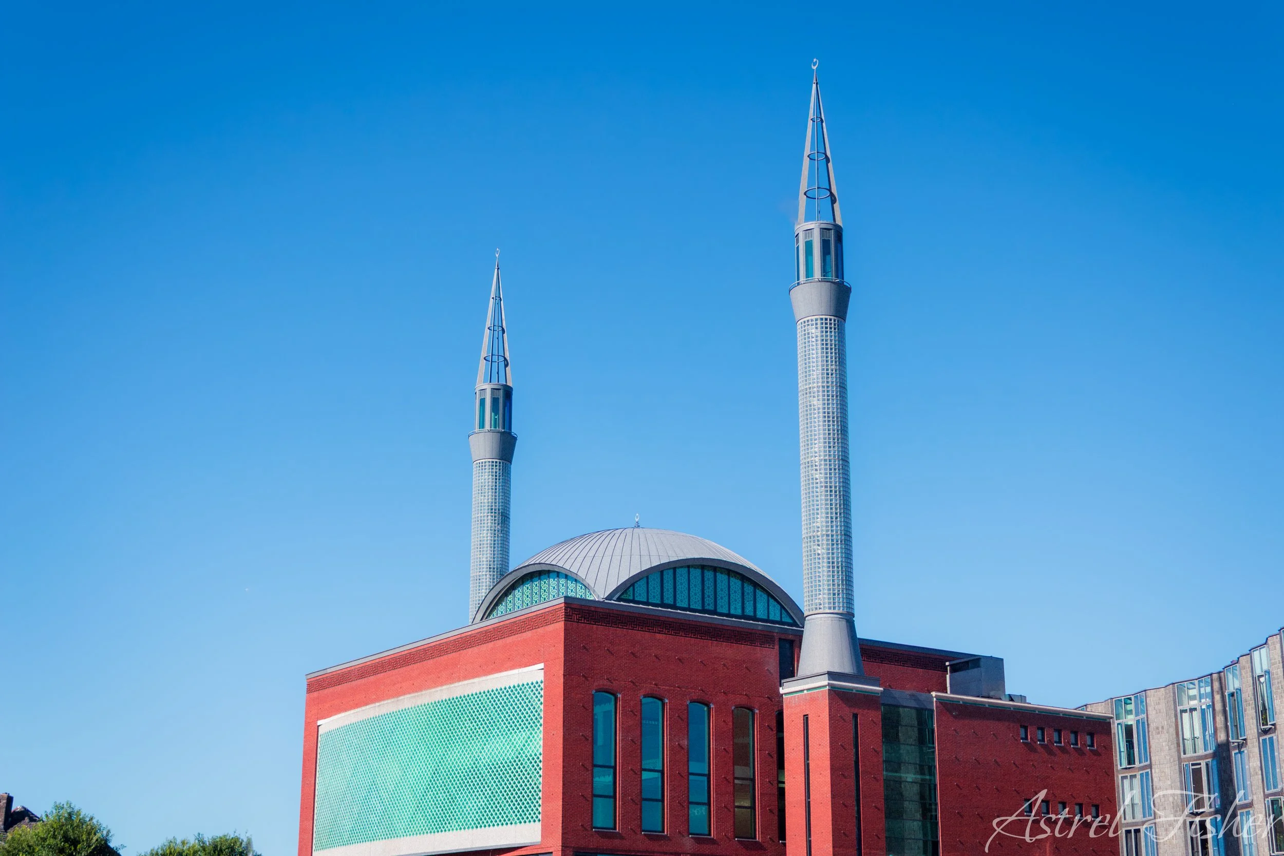 A building with two tall minarets and a dome, featuring a modern architectural design with a red brick exterior and large arched windows, set against a clear blue sky.