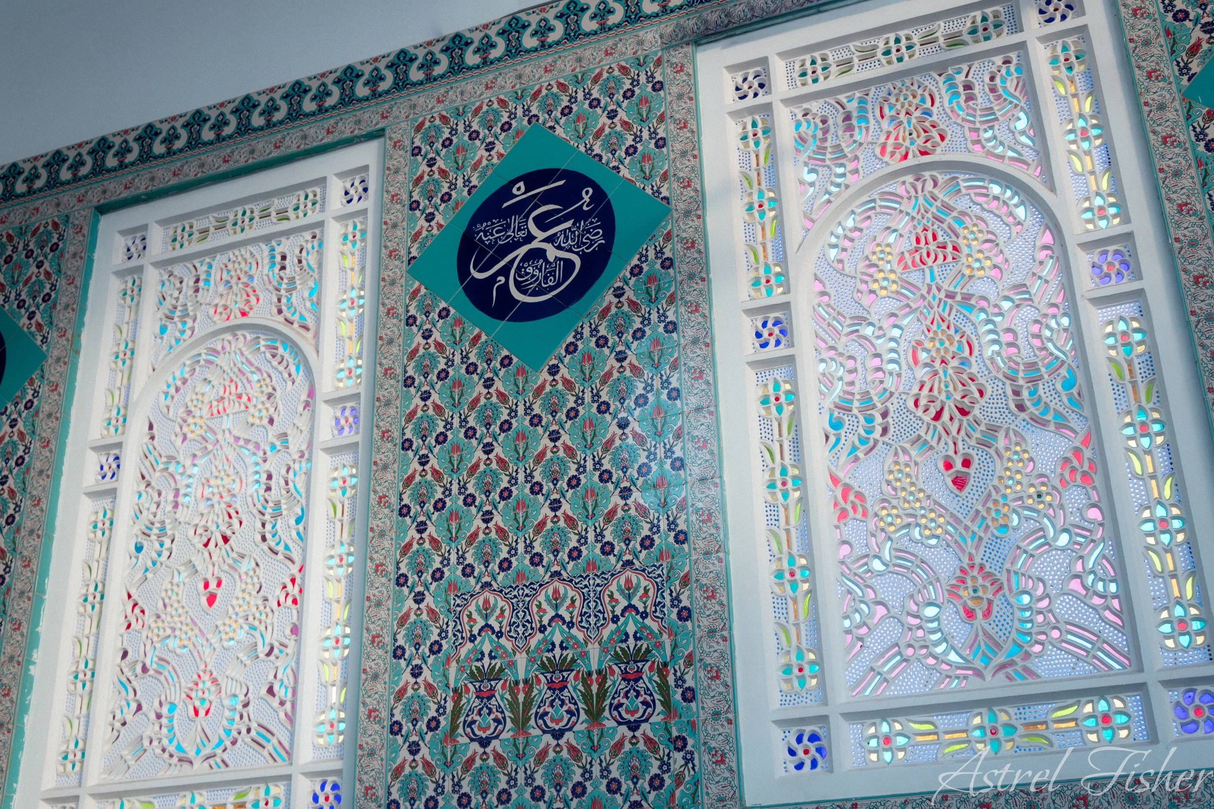 Decorative wall with intricate floral and geometric designs, stained glass windows, and an Islamic calligraphy plaque.
