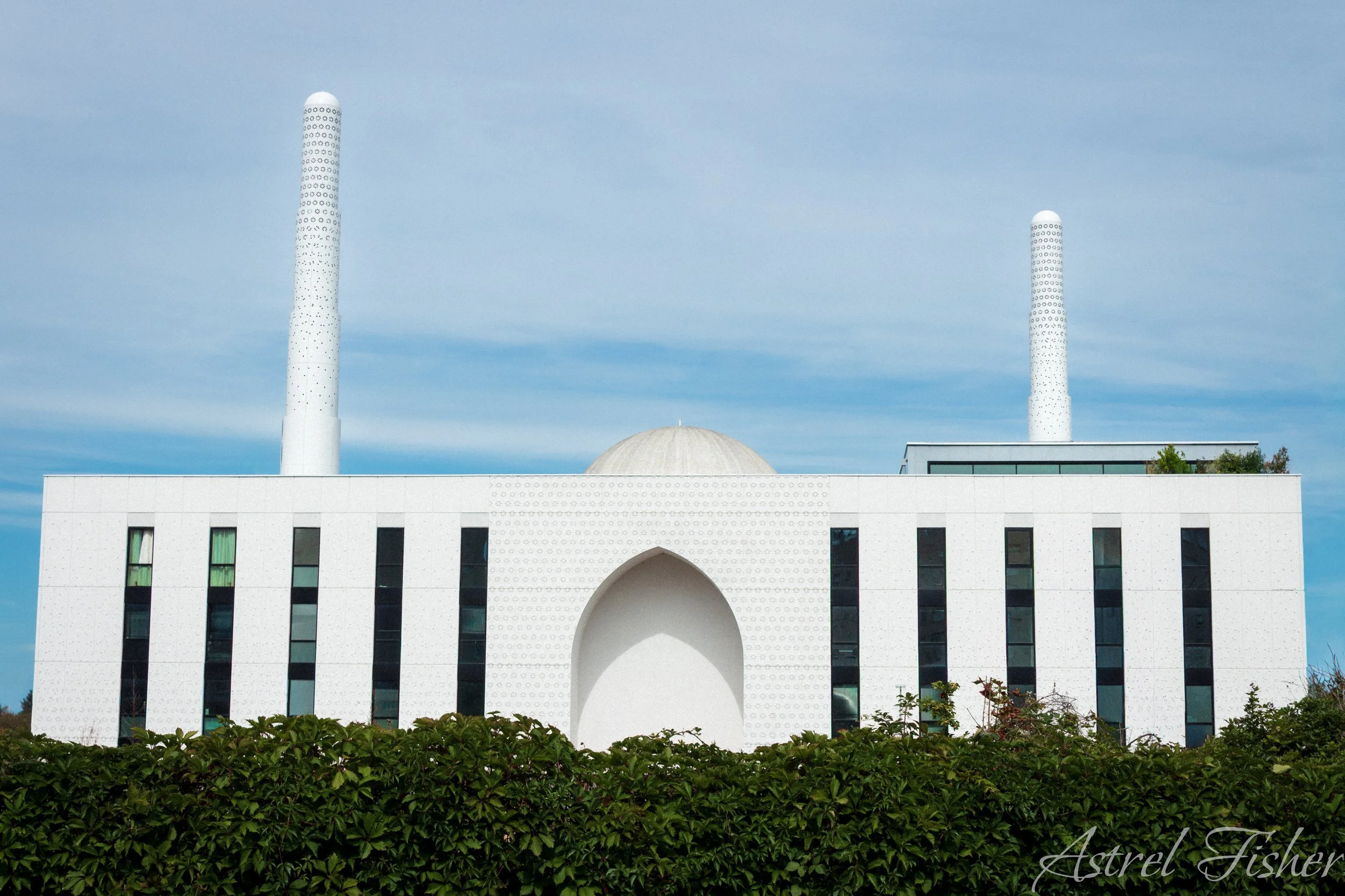 A modern white building with black windowpanes, featuring two tall white smokestacks and a dome in the center, under a partly cloudy blue sky, with greenery at the bottom.