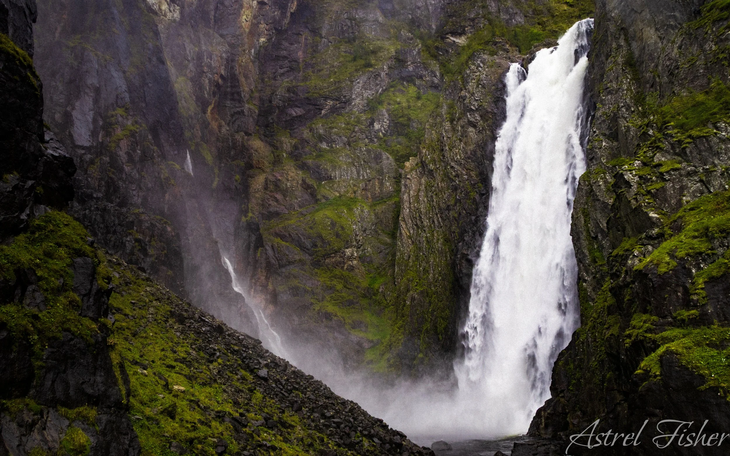 A tall waterfall cascading down a rocky cliff surrounded by green moss and vegetation, with mist rising at the base.