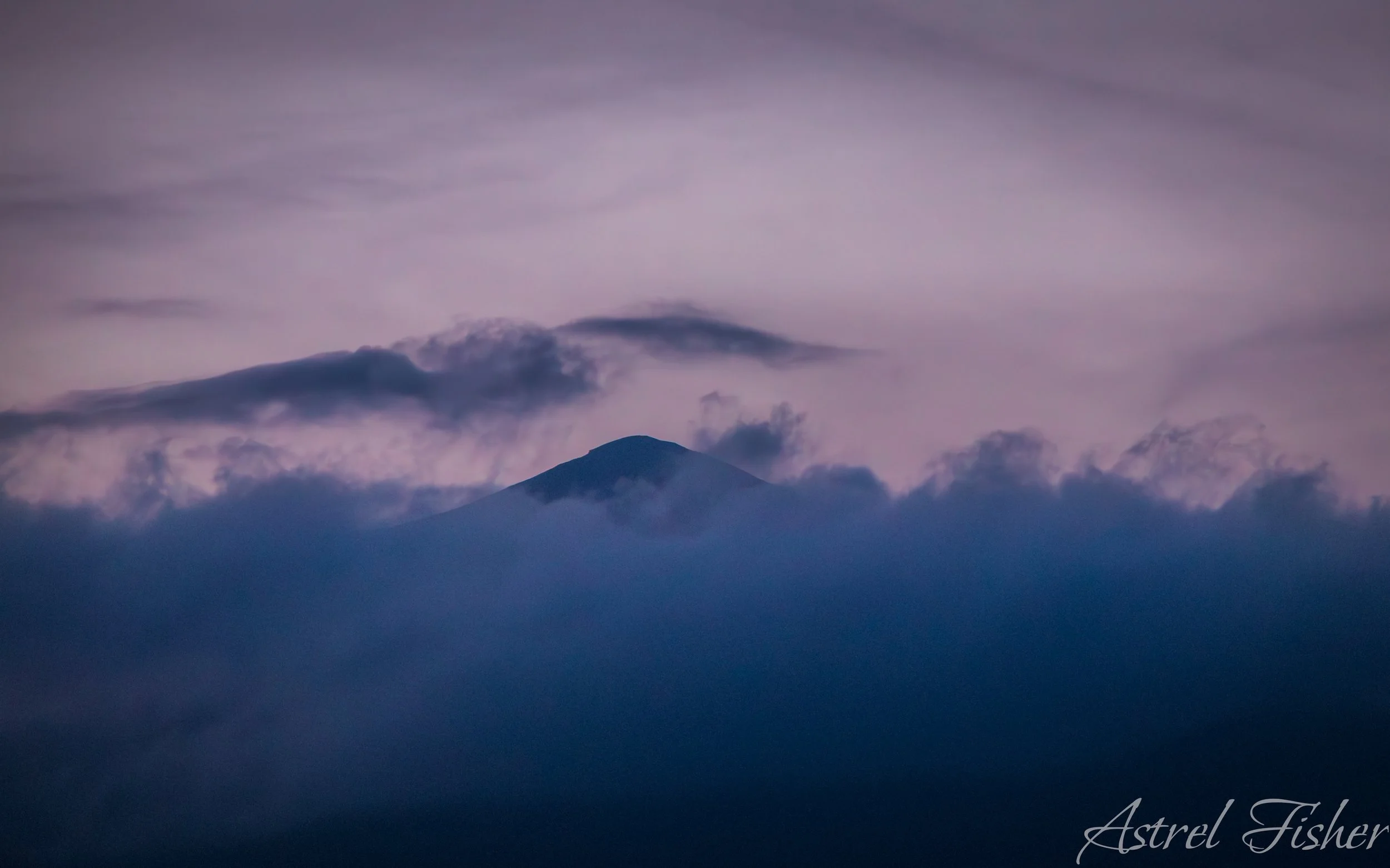 A mountain peak emerging above clouds during dusk with a purple sky.