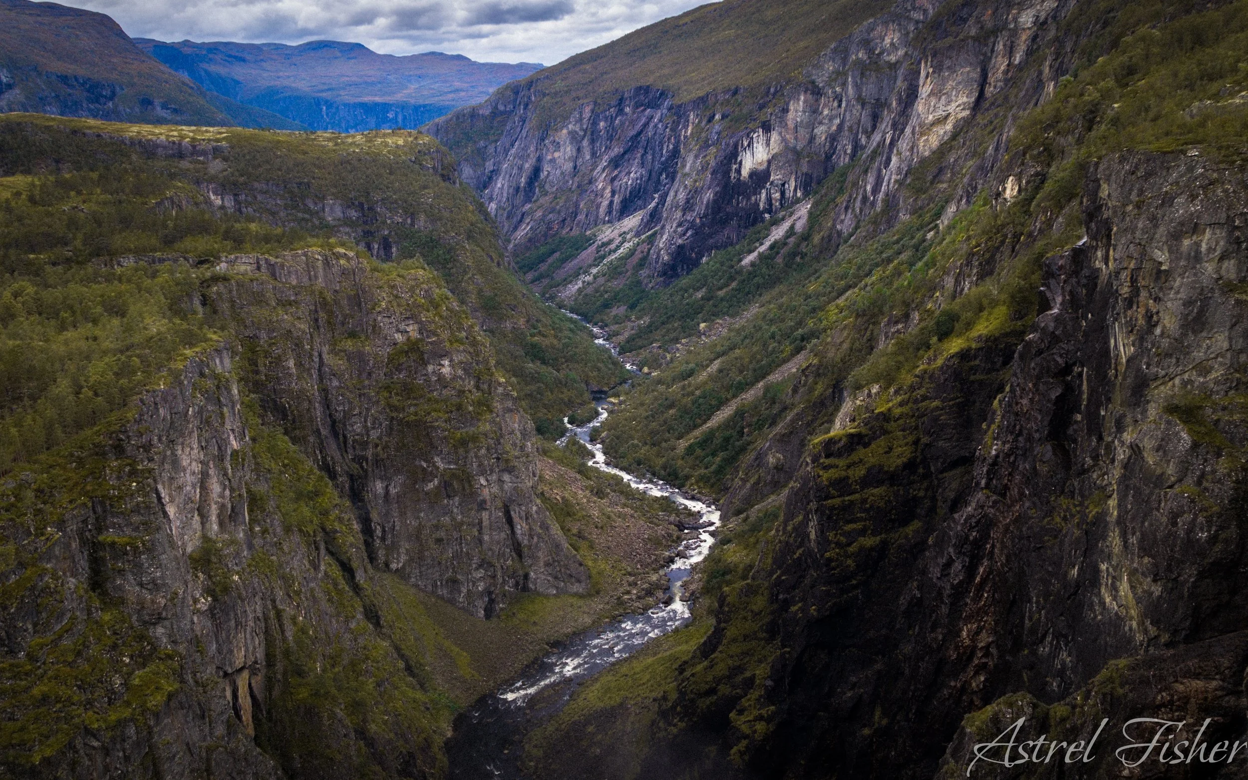 A deep, narrow canyon with steep rocky cliffs on both sides, a flowing river at the bottom, and distant mountains in the background under cloudy skies.