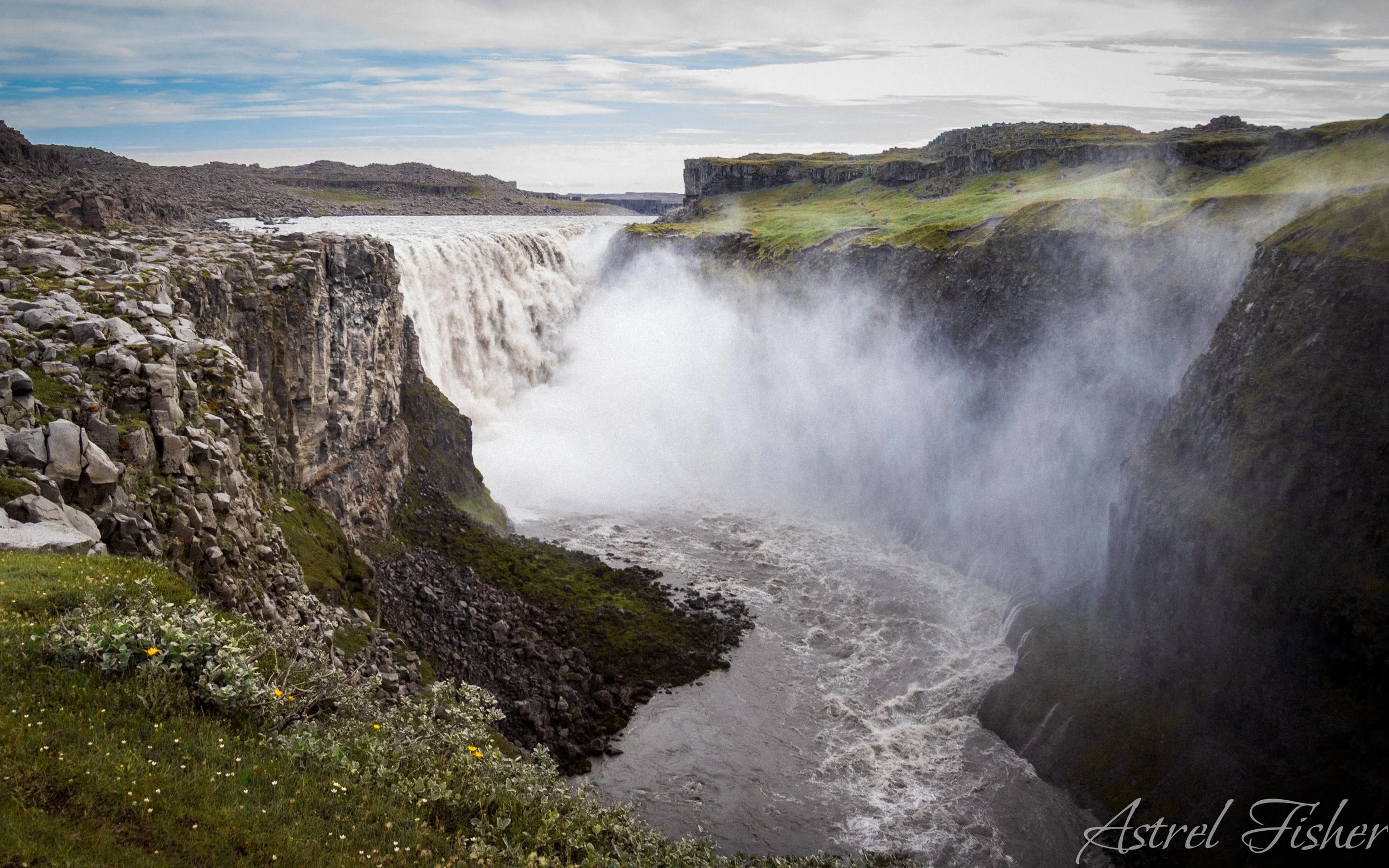 A wide waterfall flowing through a rocky canyon with green moss-covered cliffs and cloudy sky.