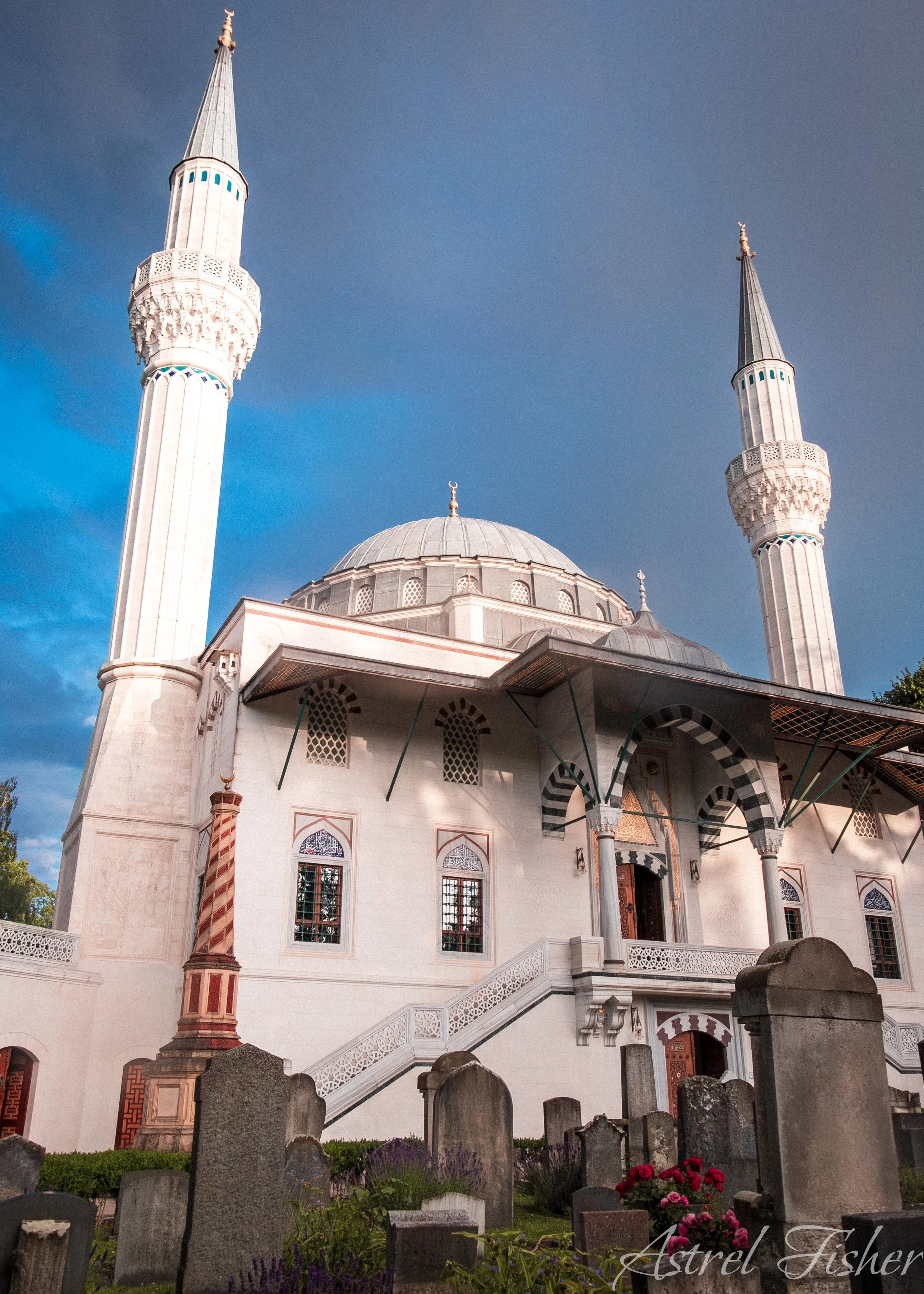A mosque with two minarets and a central dome, surrounded by a graveyard with headstones and flowers, under a partly cloudy sky.