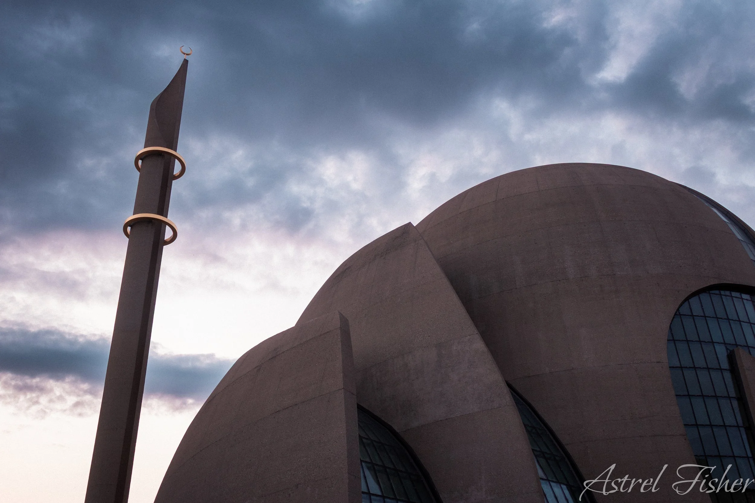 A modern, concrete architectural building with large curved surfaces and circular windows, building against a cloudy sky.