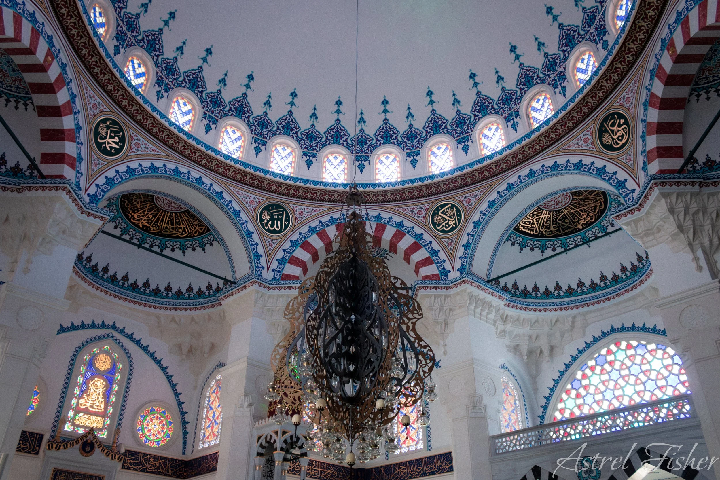 The interior of a mosque showing a decorated ceiling with intricate blue, red, and white patterns, stained glass windows, a large ornate chandelier hanging from the ceiling, and Arabic calligraphy medallions on the walls.