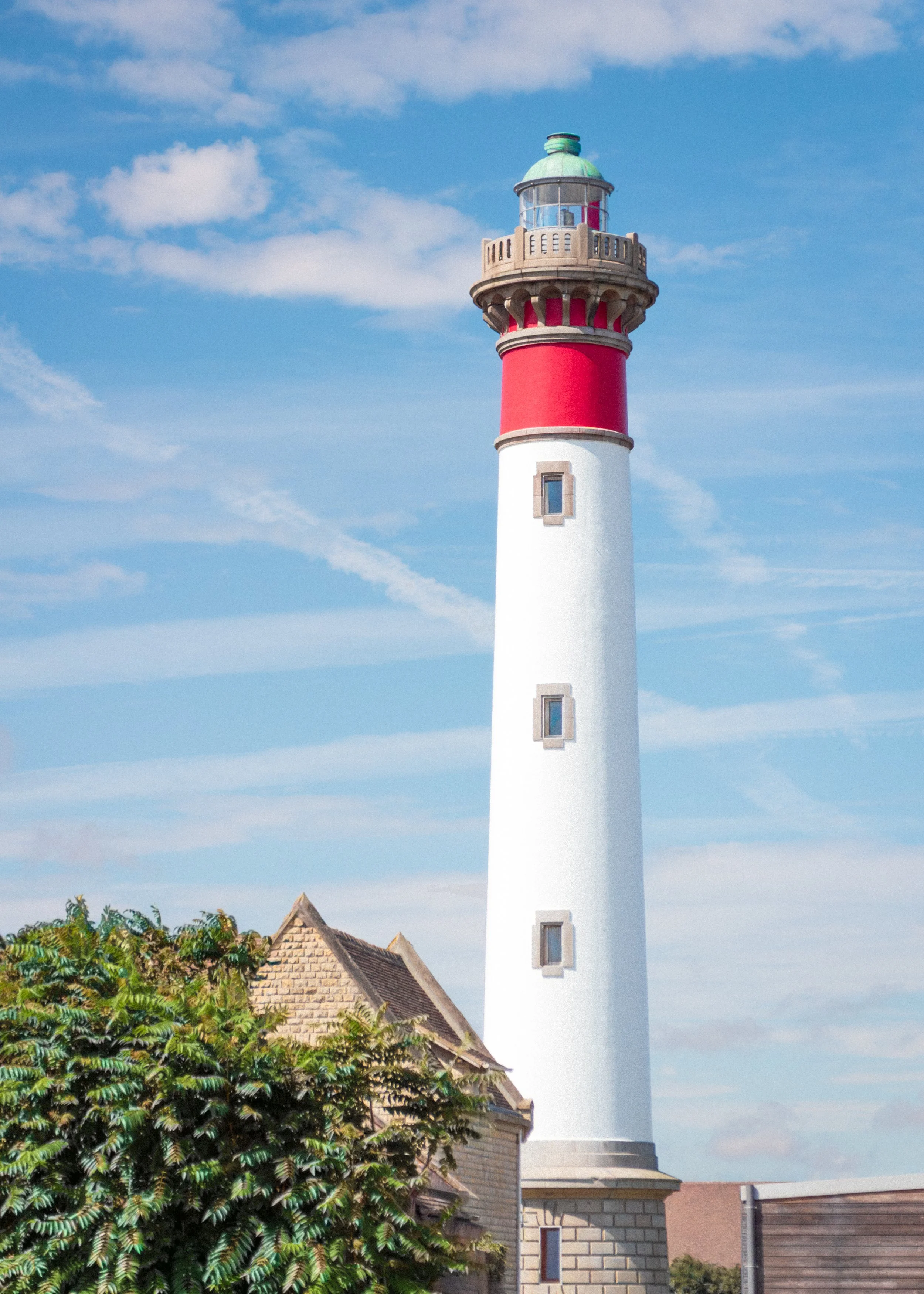 A tall white lighthouse with a red band near the top, topped with a greenish lantern room, against a blue sky with wispy clouds. There are trees and a building in the foreground.