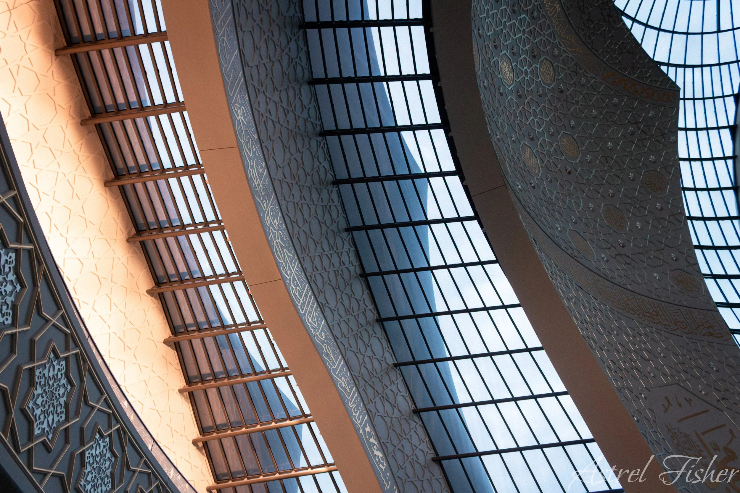Interior view of a mosque showcasing intricate Islamic geometric patterns and calligraphy on the walls and ceiling, with large windows allowing natural light to illuminate the space.