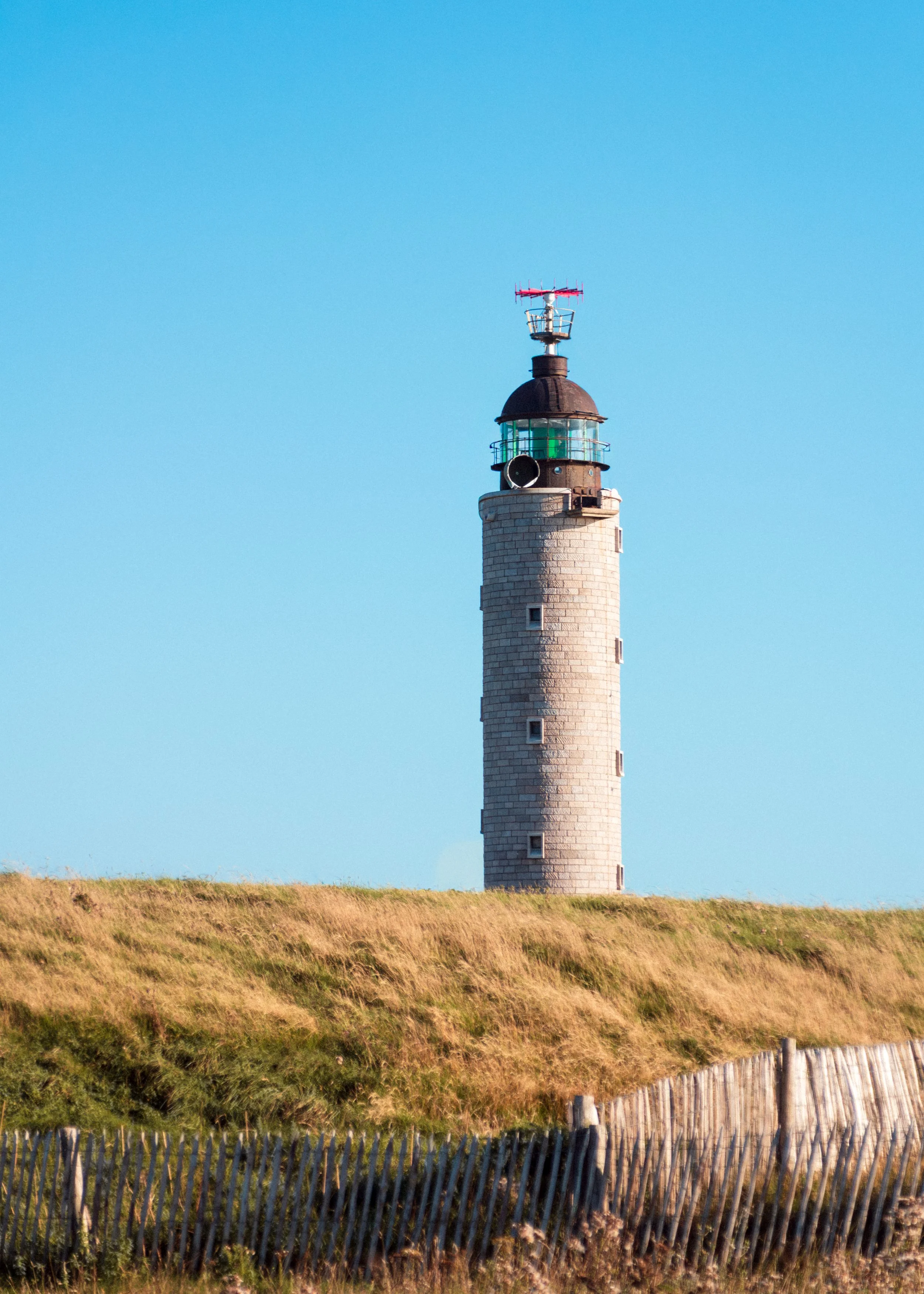 A tall stone lighthouse standing on a grassy hill with a blue sky in the background.