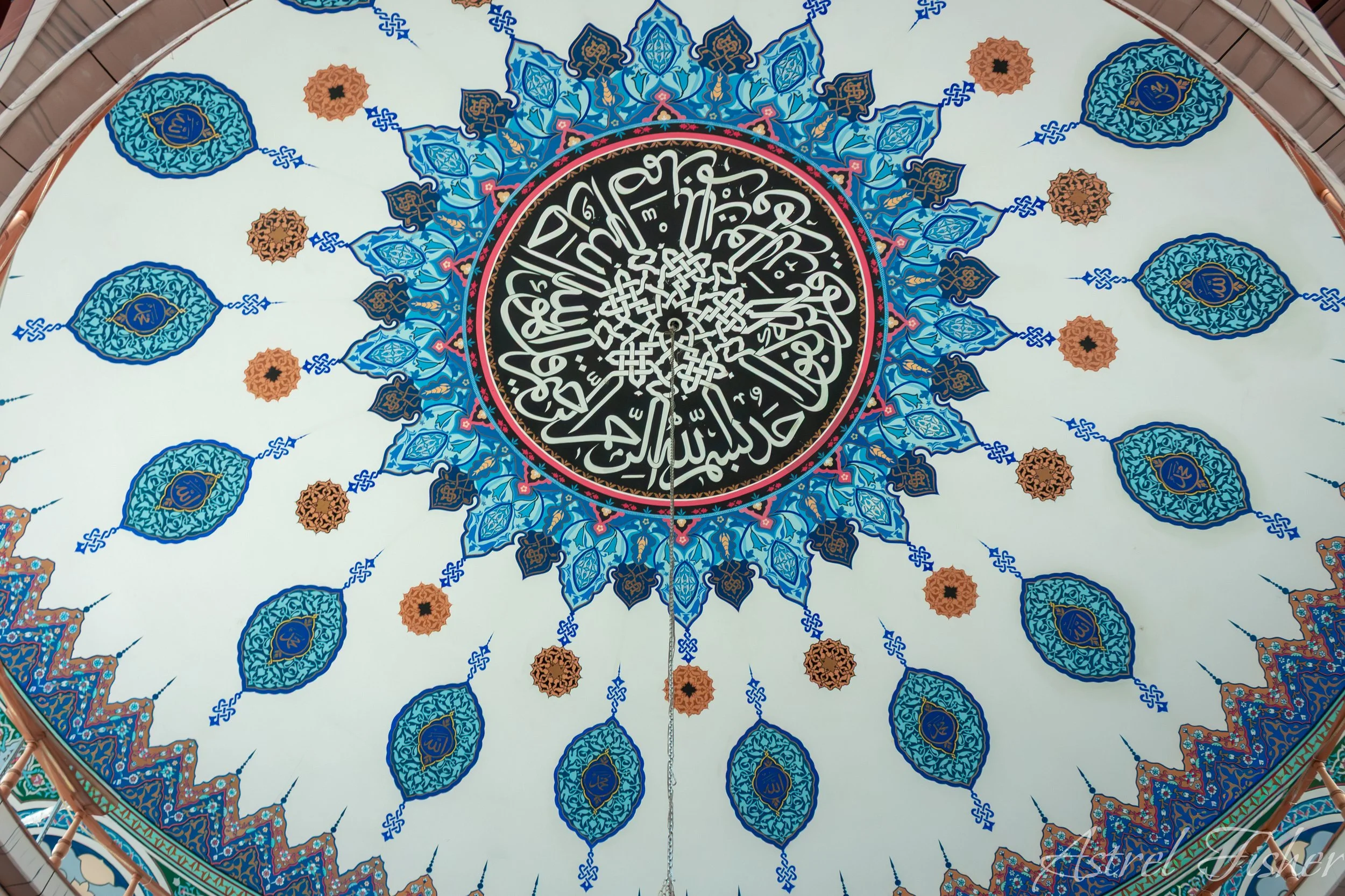 Decorative ceiling with intricate blue, black, pink, and gold Islamic calligraphy and geometric patterns, featuring a central black circle with white Arabic script.