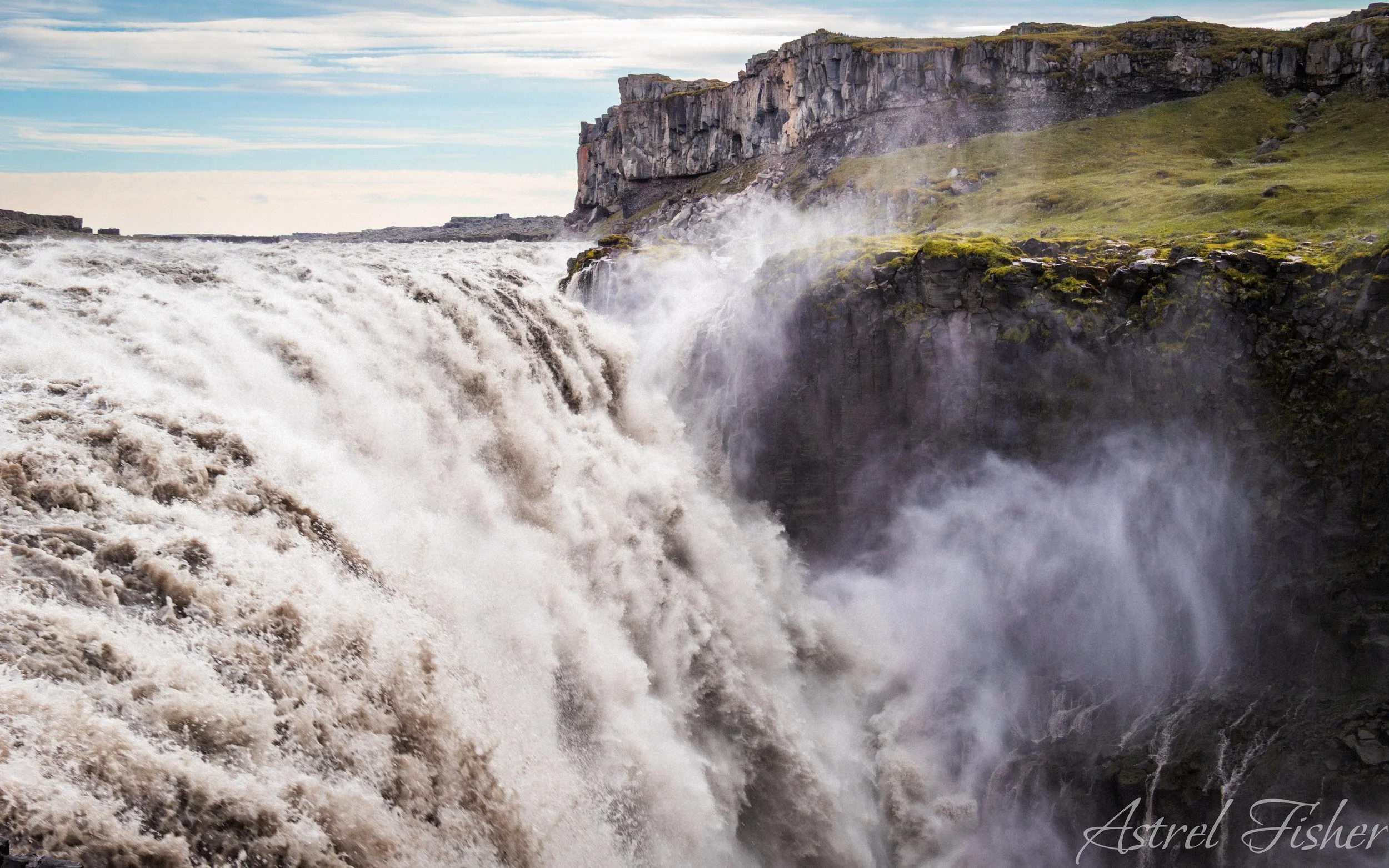 A powerful waterfall cascading down a rocky cliff with foamy water and mist rising, surrounded by green moss and grass under a cloudy sky.