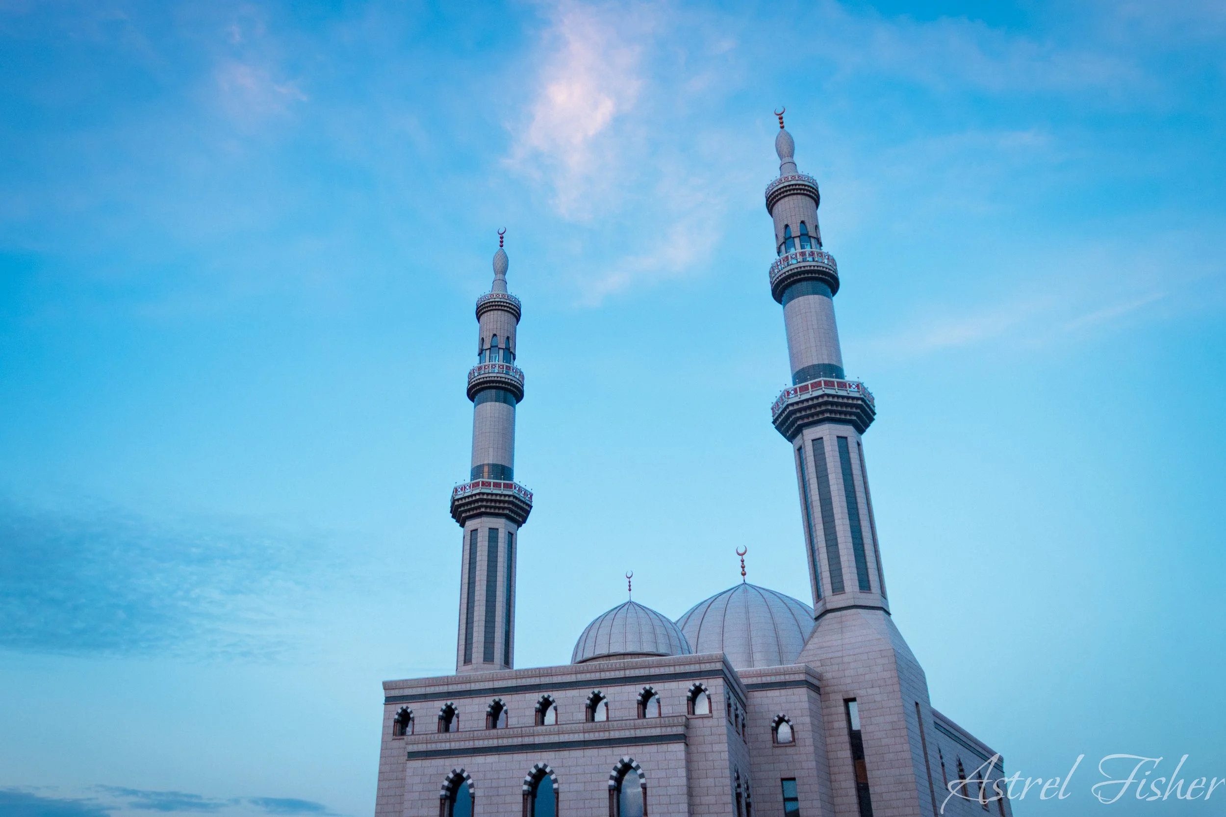 A mosque with two tall minarets with crescent moons on top, against a blue sky with scattered clouds.