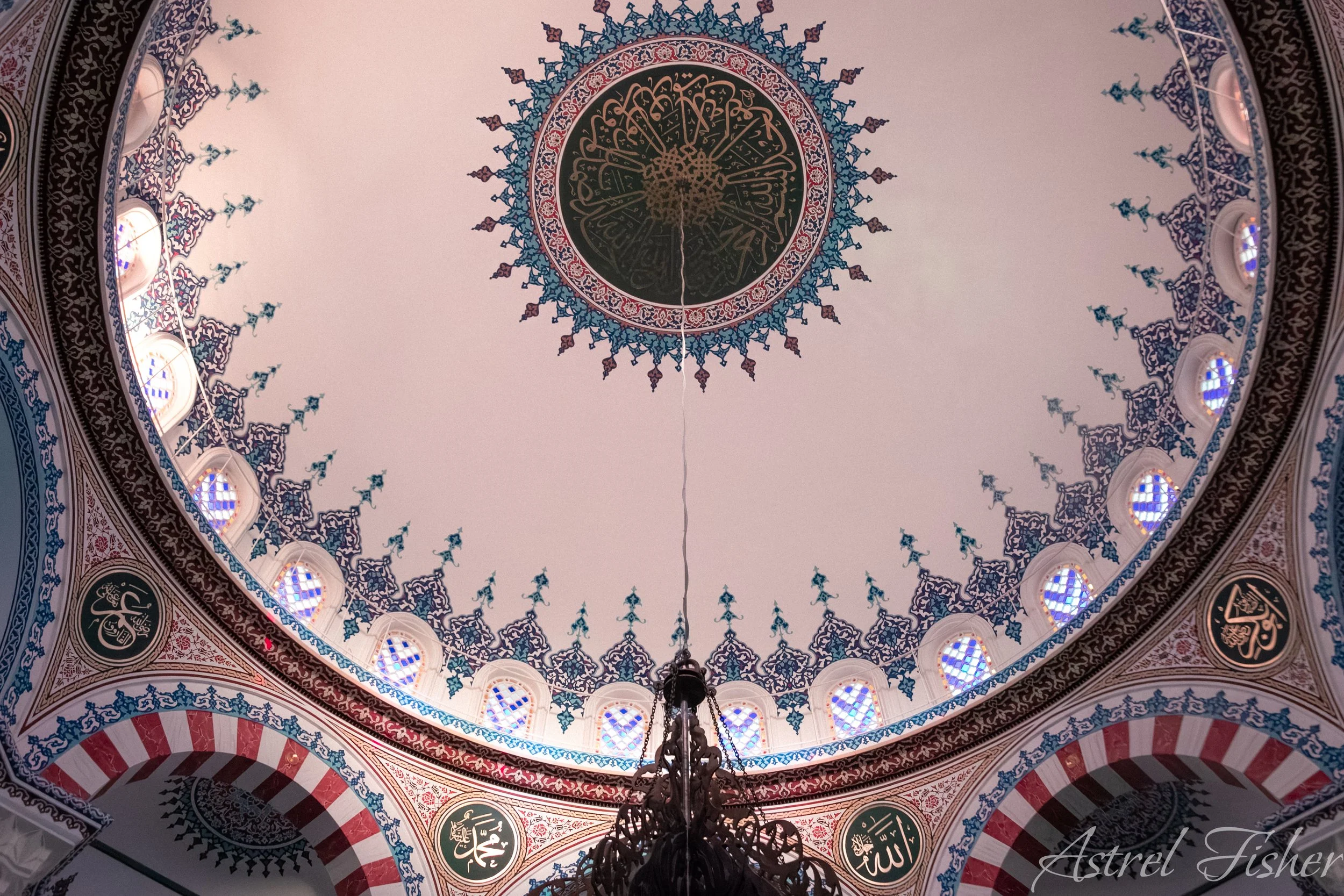 Interior view of an ornately decorated Islamic mosque dome with intricate blue, red, and gold patterns, Arabic calligraphy, and stained glass windows.