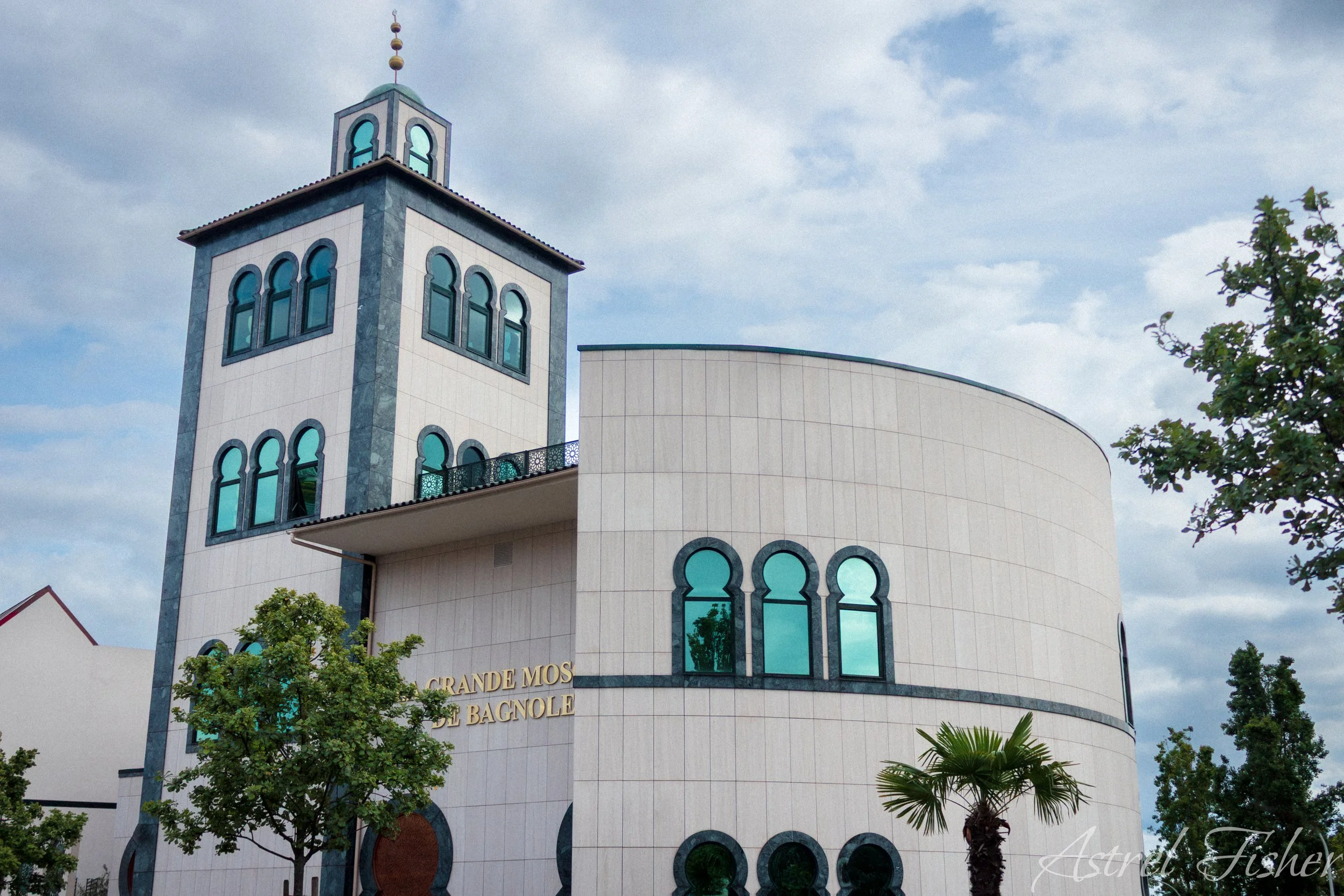 A modern building with a prominent tower featuring arched windows and decorative elements, with the name 'Grandes Musées de Bagnole' on the facade, surrounded by green trees under a partly cloudy sky.