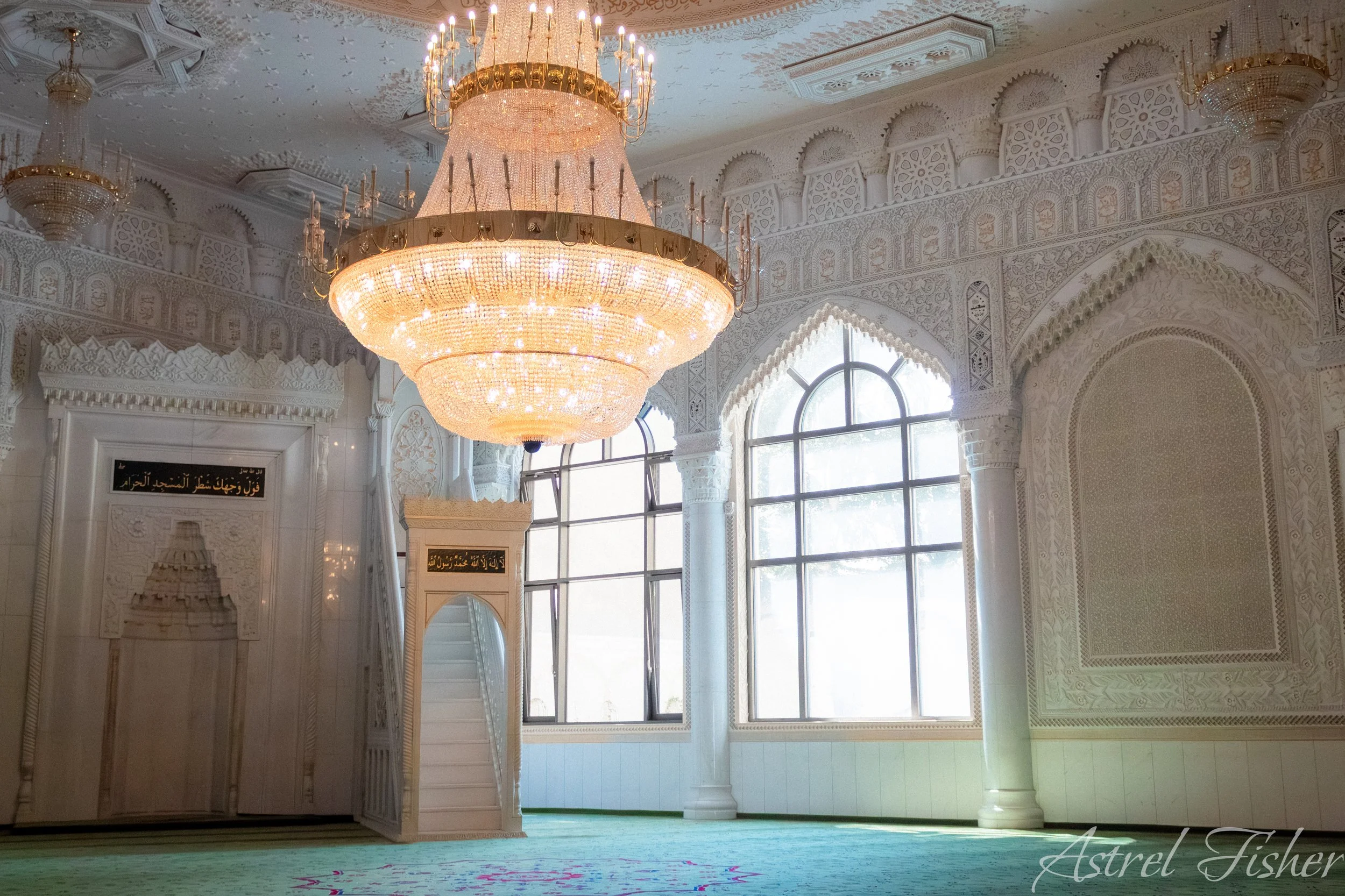 An ornate interior with intricate white carvings on the walls and ceiling, large arched windows, and a grand chandelier hanging from the ceiling.