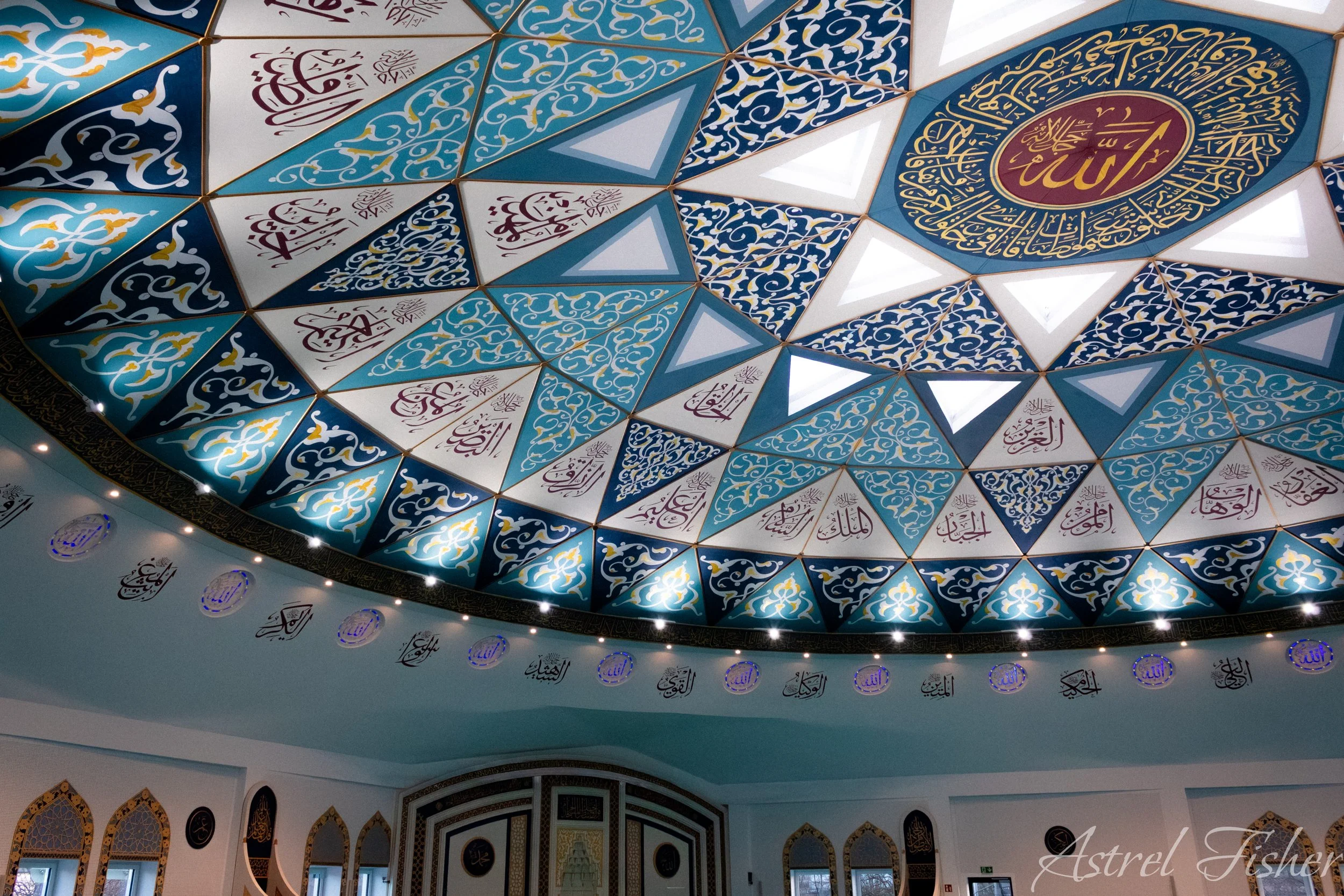Interior view of a mosque ceiling with intricate geometric patterns and Arabic calligraphy in blue, gold, and white colors.