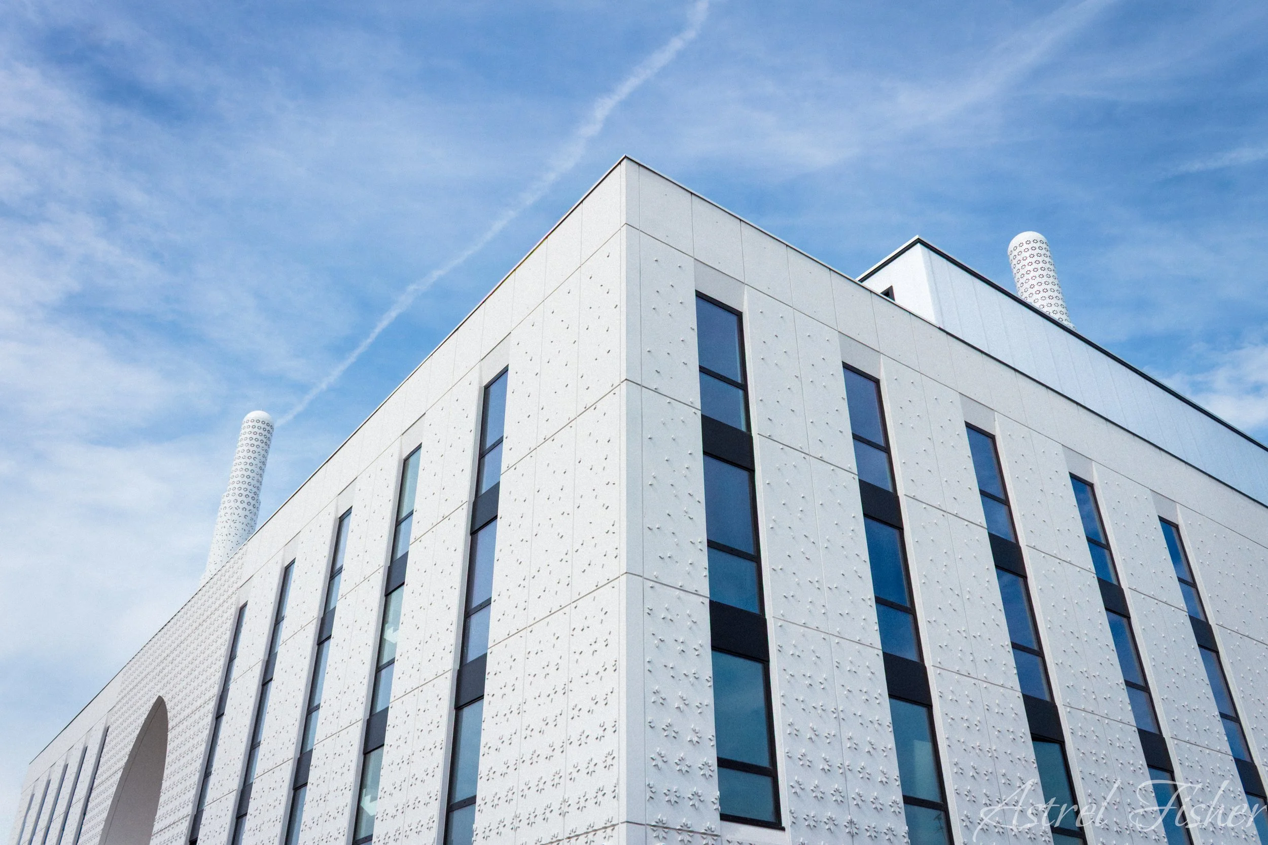 The image shows the corner of a modern white building with multiple rectangular windows, set against a blue sky with wispy clouds and a contrail. There are two white cylindrical structures with circular cut-out patterns on the roof.