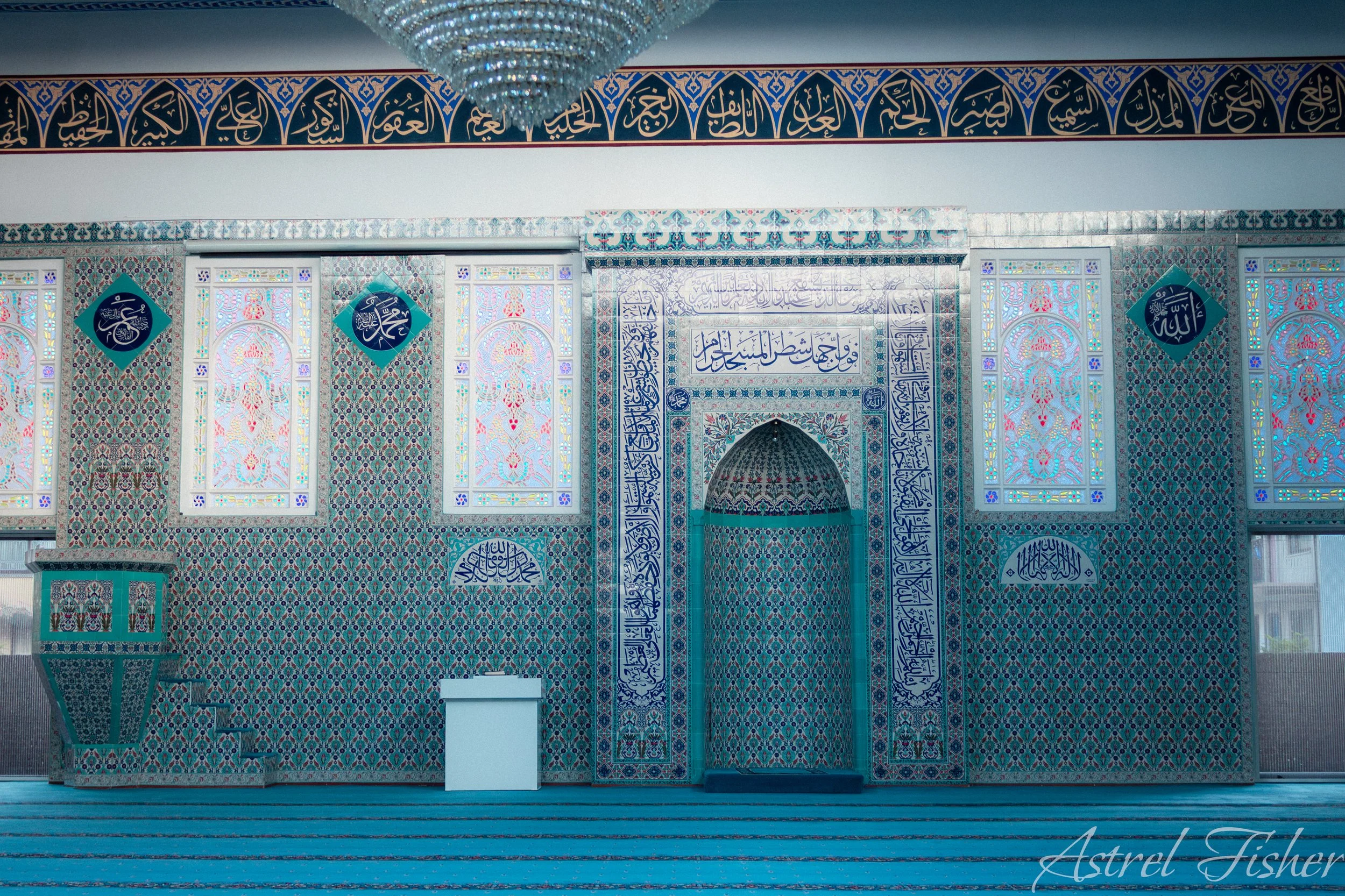 Interior of a mosque with intricate blue and white tile designs, stained glass windows, and a chandelier hanging from the ceiling.