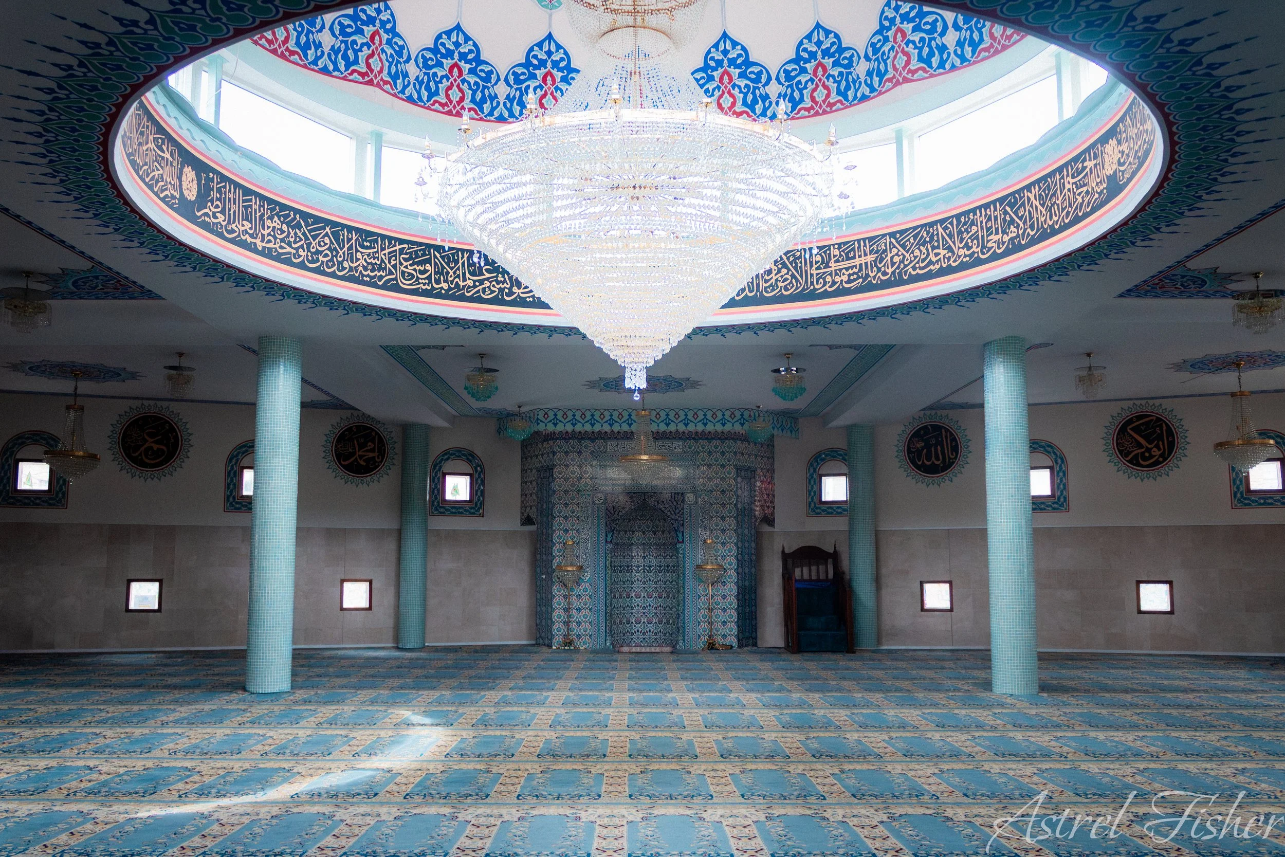Interior of a mosque with ornate blue and gold decorations, a large chandelier, columns covered in light blue tiles, and Arabic calligraphy around the upper walls.