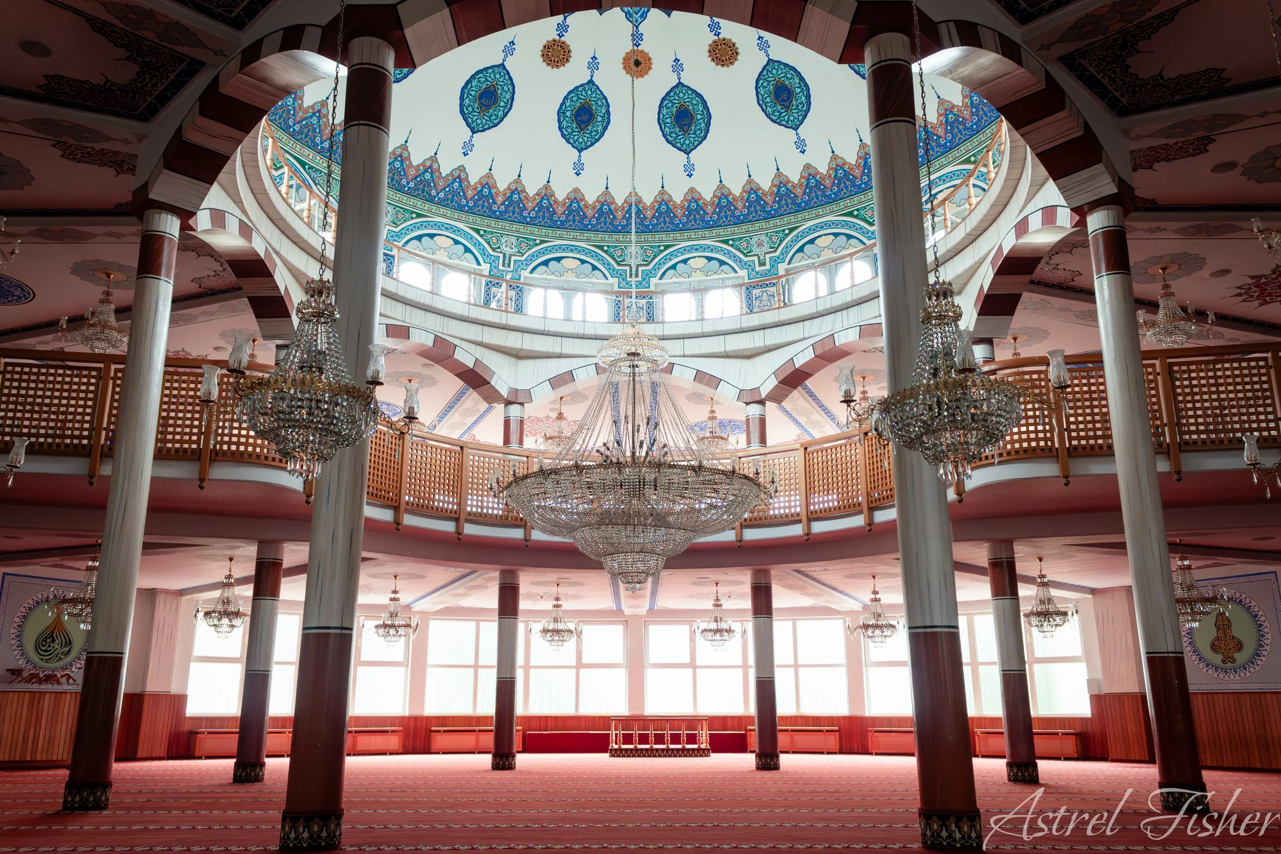 Interior of a mosque with a large central chandelier, ornate decor, decorative ceiling with Islamic motifs, and tall white columns supporting a balcony with wooden railings, illuminated by natural light from large windows.