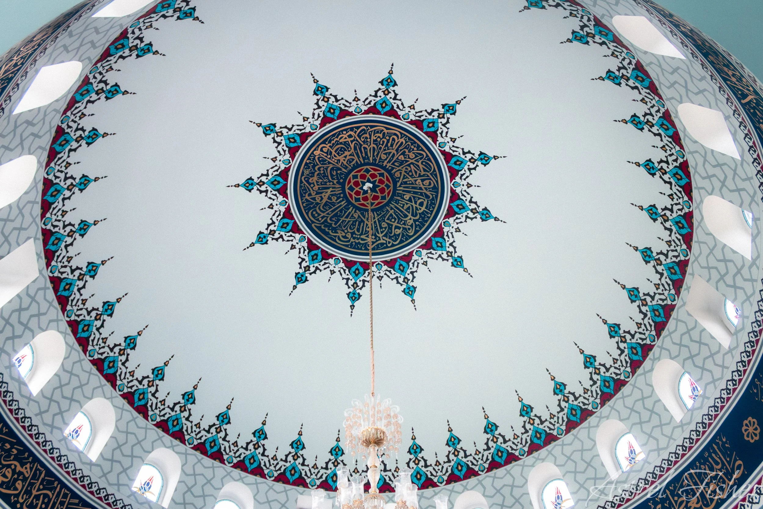The interior of an Islamic mosque dome, decorated with intricate geometric and floral patterns in blue, gold, and red, with a chandelier hanging from the center.