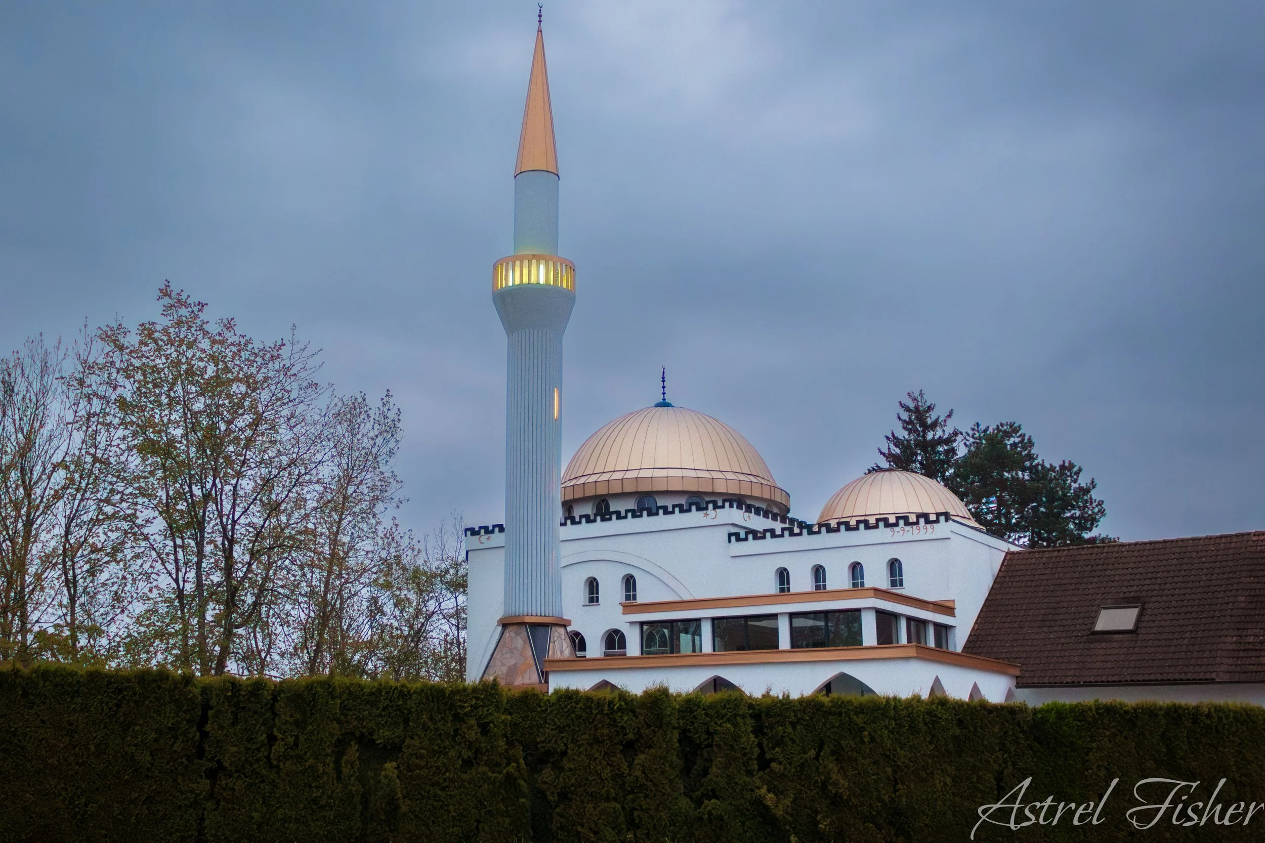 A mosque with a large central dome and a tall minaret with a golden spire, surrounded by trees and a hedge, under a cloudy sky at dusk.