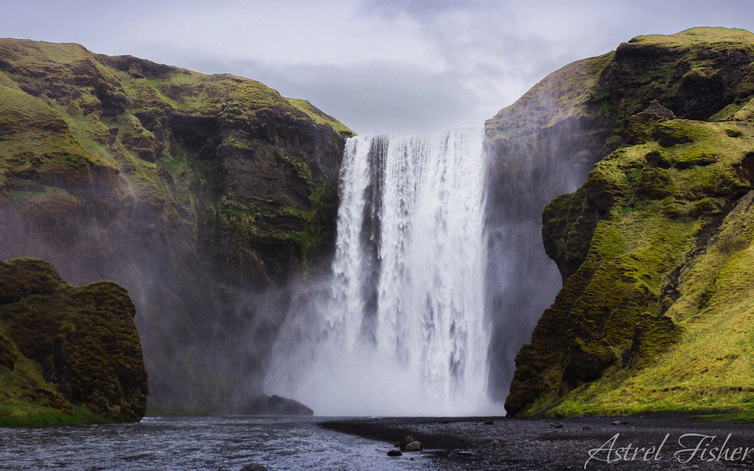 A scenic view of a waterfall cascading between green, moss-covered cliffs with a river in the foreground.