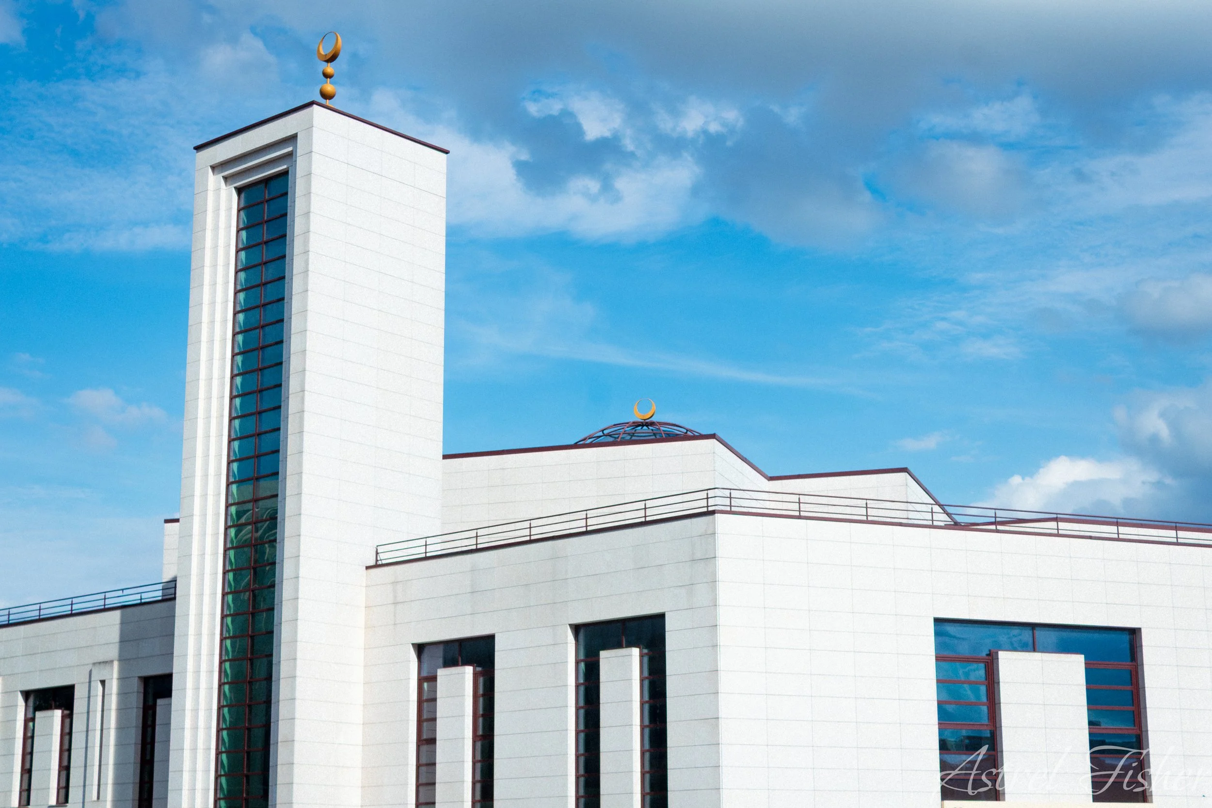 White modern building with glass windows and balconies under a partly cloudy blue sky, topped with a moon and star symbol.