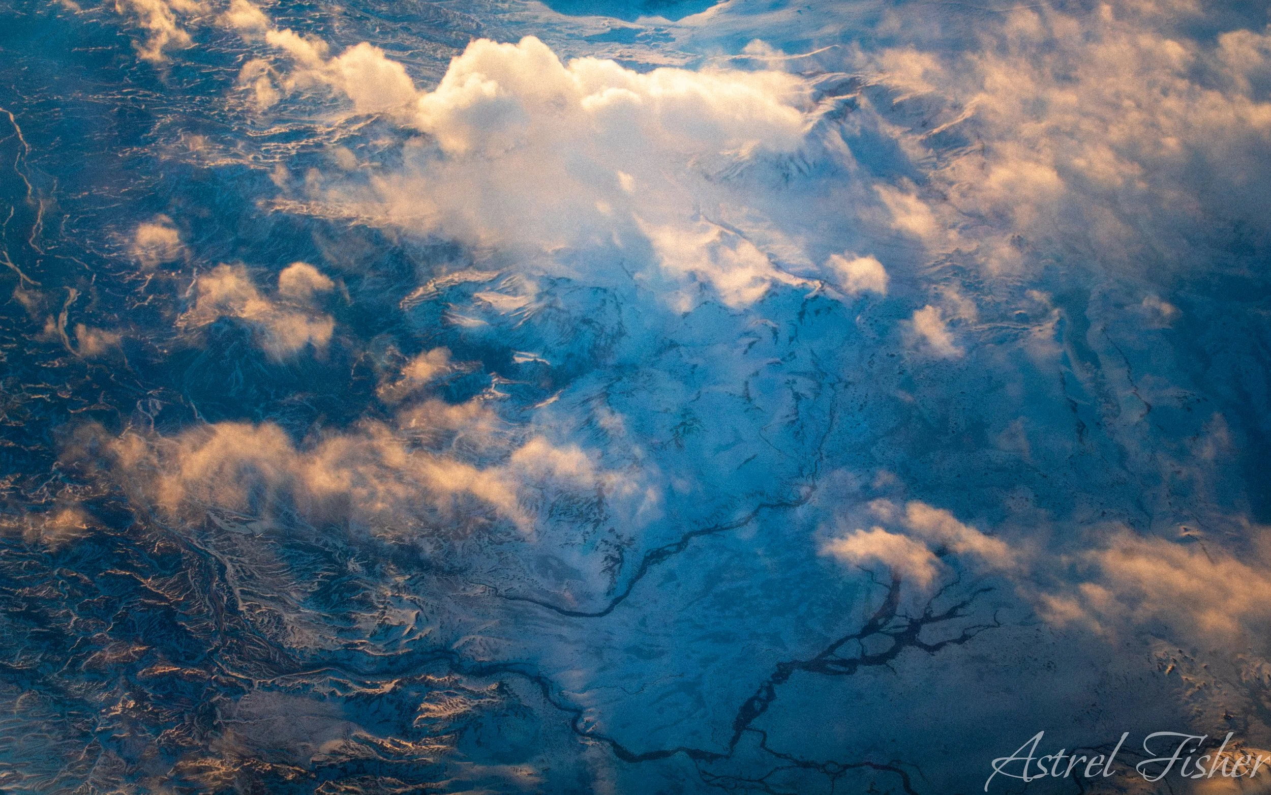 Aerial view of mountain peaks and valleys covered in snow, with clouds overhead, during sunset or sunrise.