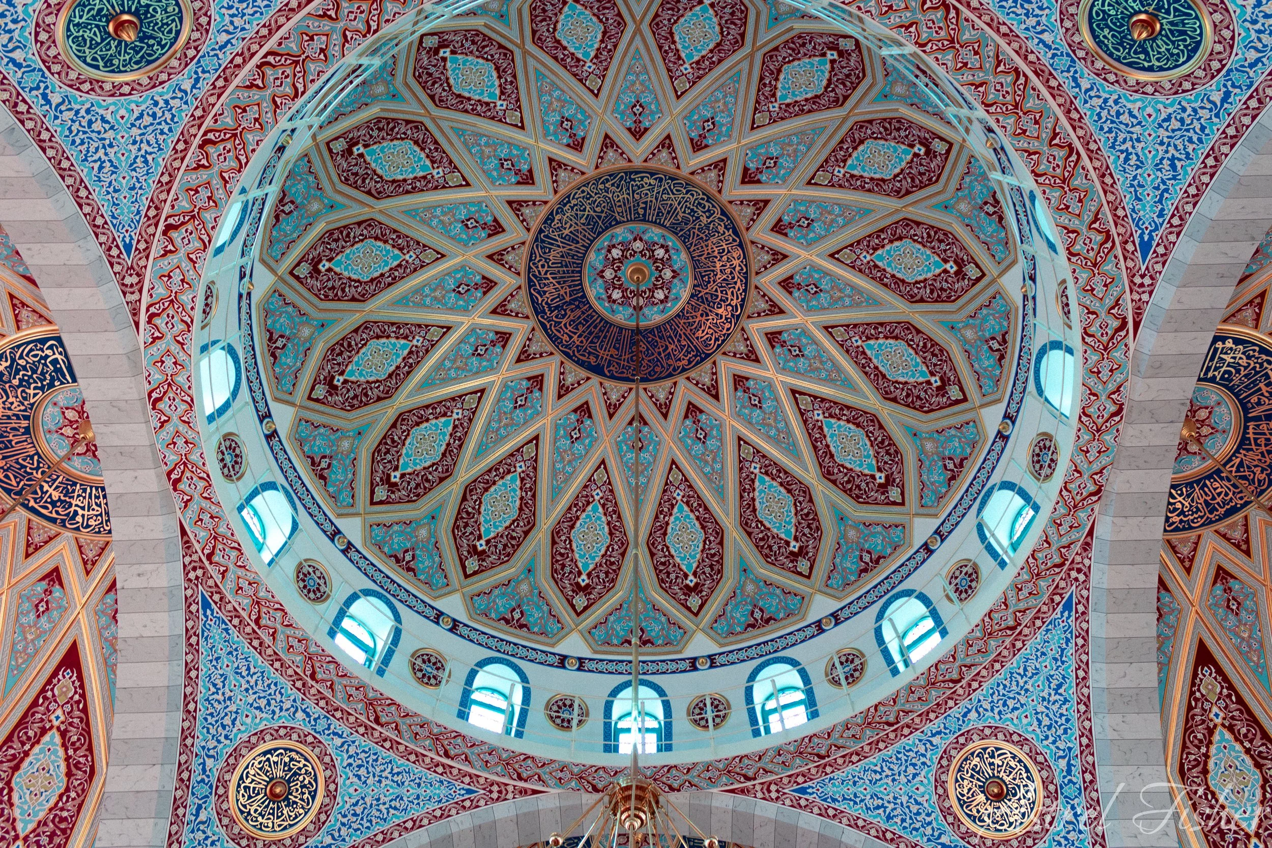 Intricate interior ceiling of a mosque featuring a large central dome with detailed geometric and floral patterns in red, blue, and gold colors, surrounded by smaller decorative medallions and arched windows.
